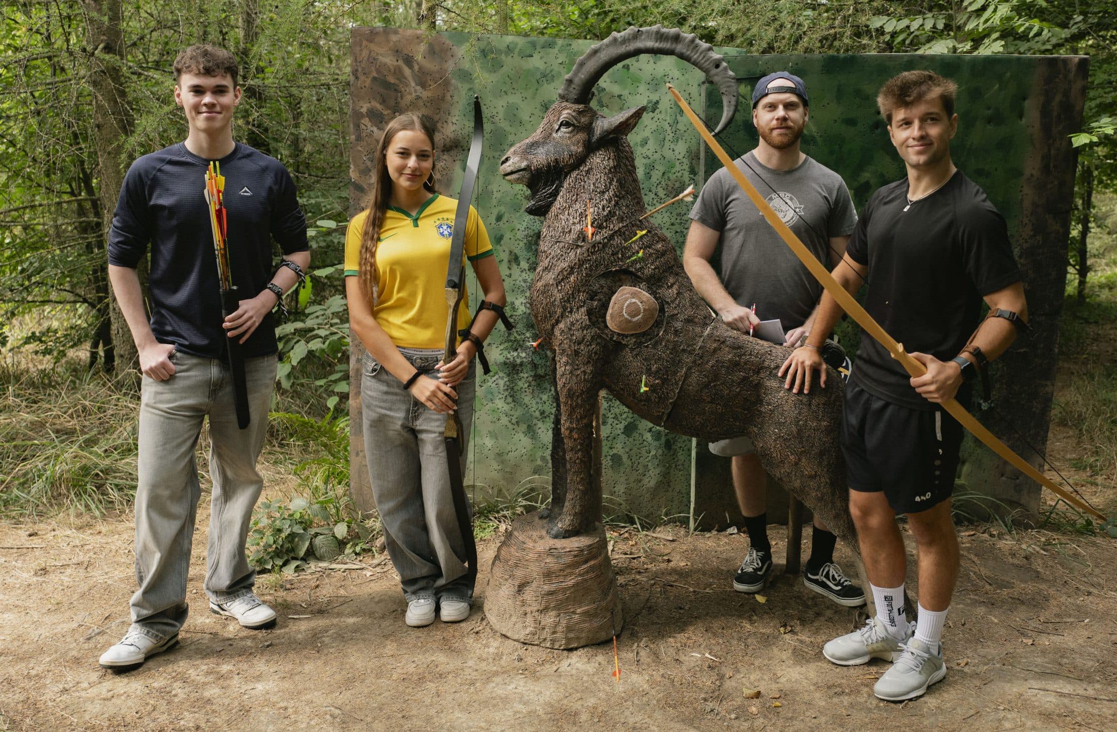 Vier junge Erwachsene stehen in einem Wald neben einer lebensgroßen Bogenschießscheibe in Form einer Bergziege. Jeder von ihnen hält einen Bogen und trägt einen Köcher, posiert und lächelt in die Kamera. Sie sind von Bäumen und Grünflächen umgeben.