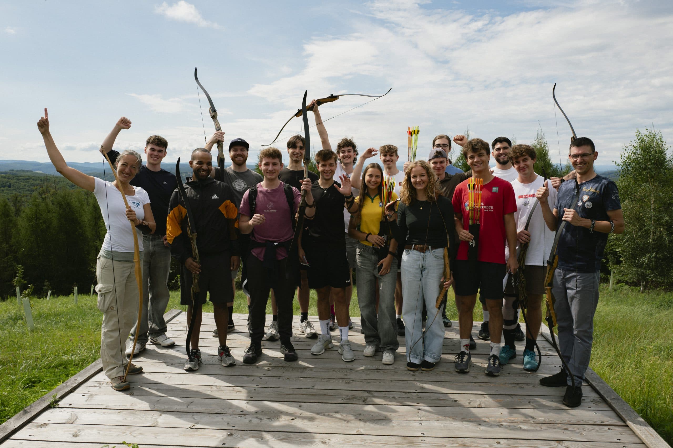 Eine Gruppe von Menschen steht im Freien auf einer hölzernen Plattform zusammen, hält Pfeil und Bogen und lächelt in die Kamera. Im Hintergrund sind Bäume und Hügel unter einem teilweise bewölkten Himmel zu sehen.