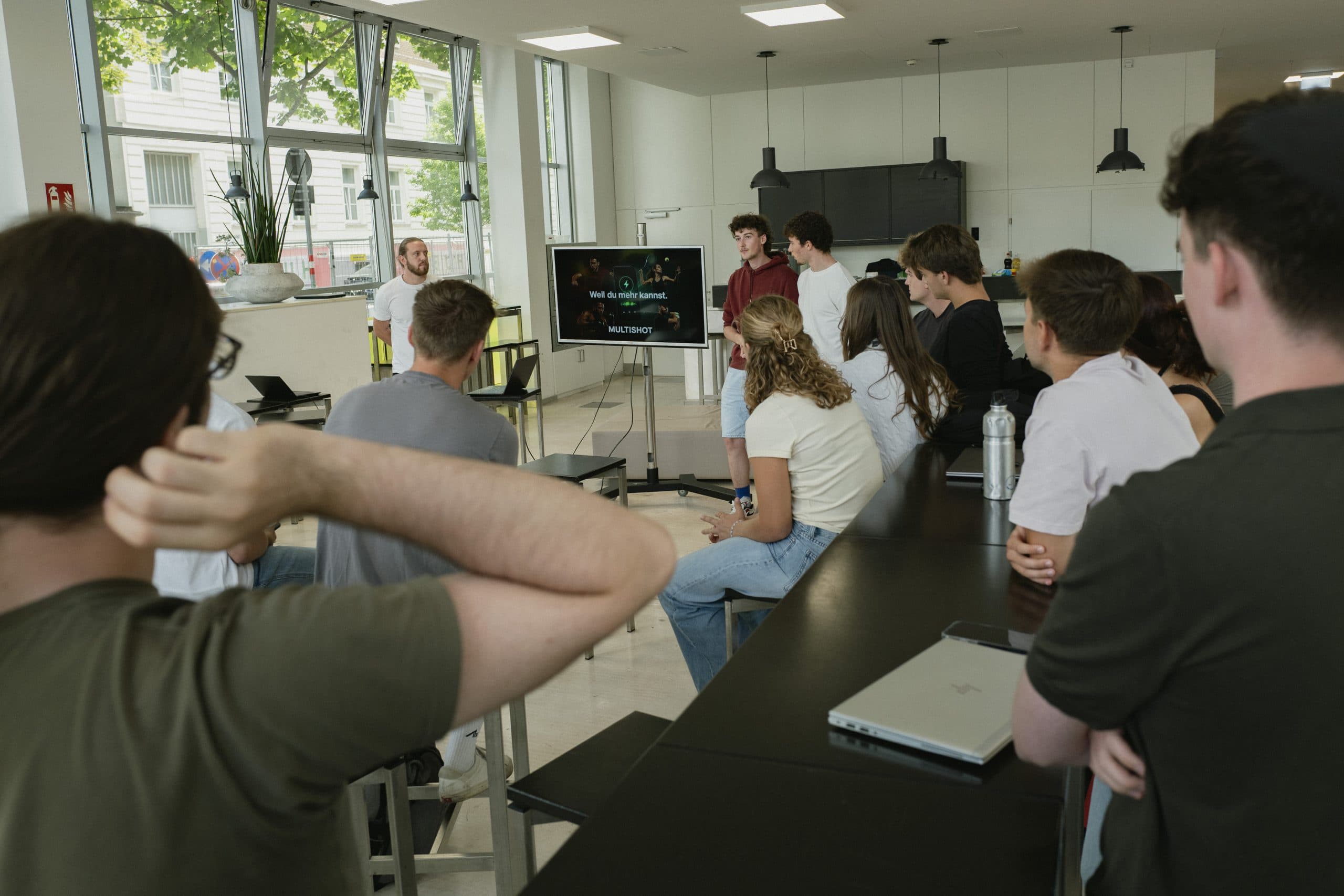 In einem modernen Klassenzimmer sitzt und steht eine Gruppe von Schülern um einen Bildschirm herum und sieht zu, wie zwei Moderatoren an der Stirnseite einen Vortrag halten; auf den Tischen stehen Laptops und Wasserflaschen.