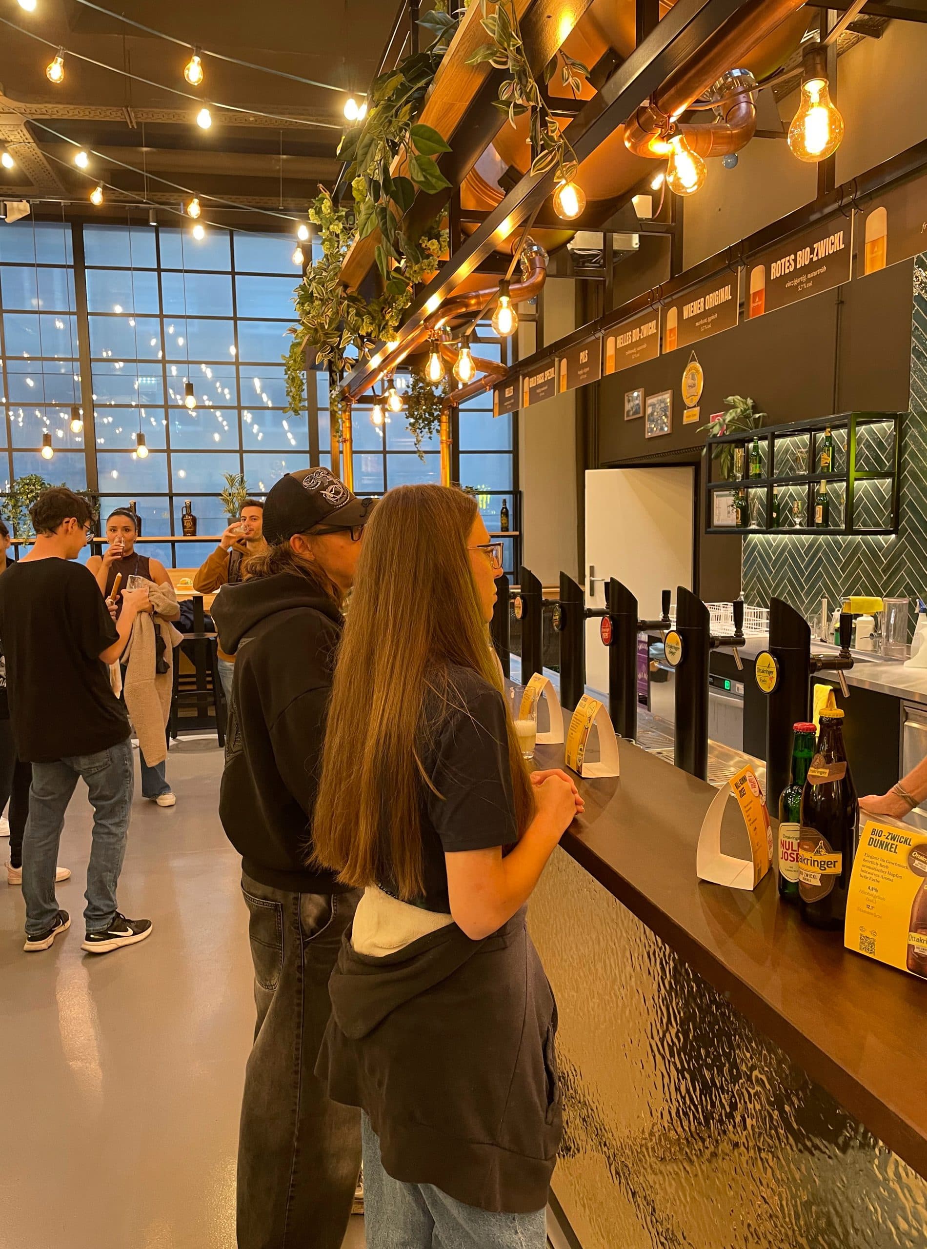 Menschen stehen an einem Tresen in einem modernen, gut beleuchteten Restaurant mit Hängelampen und großen Fenstern. Speisen und Getränke stehen auf dem Tresen, und andere warten oder unterhalten sich im Hintergrund. Die Atmosphäre ist locker und lebendig.