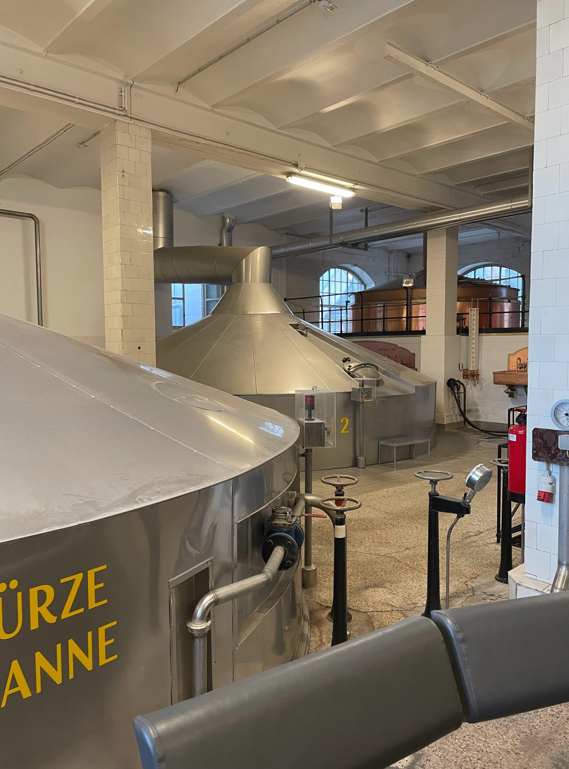 Große Metalltanks und -rohre in einer Brauerei mit weiß gekachelten Wänden, hohen Decken und natürlichem Licht, das durch gewölbte Fenster einfällt. Die Tanks sind in deutscher Sprache beschriftet.