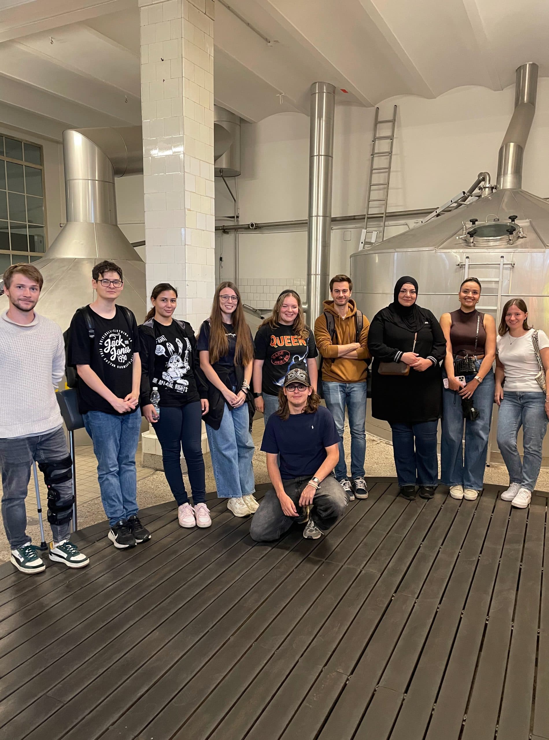 Eine Gruppe von zehn Personen posiert und lächelt in einem Innenraum vor großen Metalltanks und weiß gekachelten Säulen, die an eine Industrie- oder Brauereikulisse erinnern. Neun stehen, eine Person kniet in der vorderen Mitte.