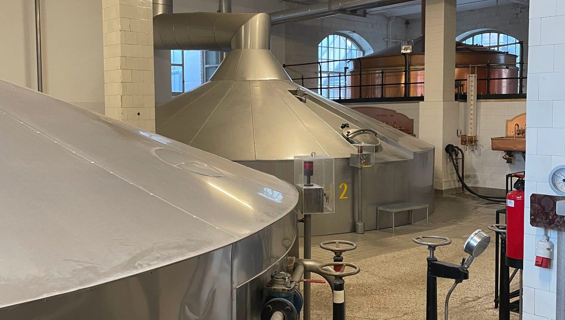 Große Edelstahltanks und -geräte in einer Brauerei, mit Rohren, Ventilen und Industriebeleuchtung. Der Raum hat weiß gekachelte Wände und große Fenster im Hintergrund.