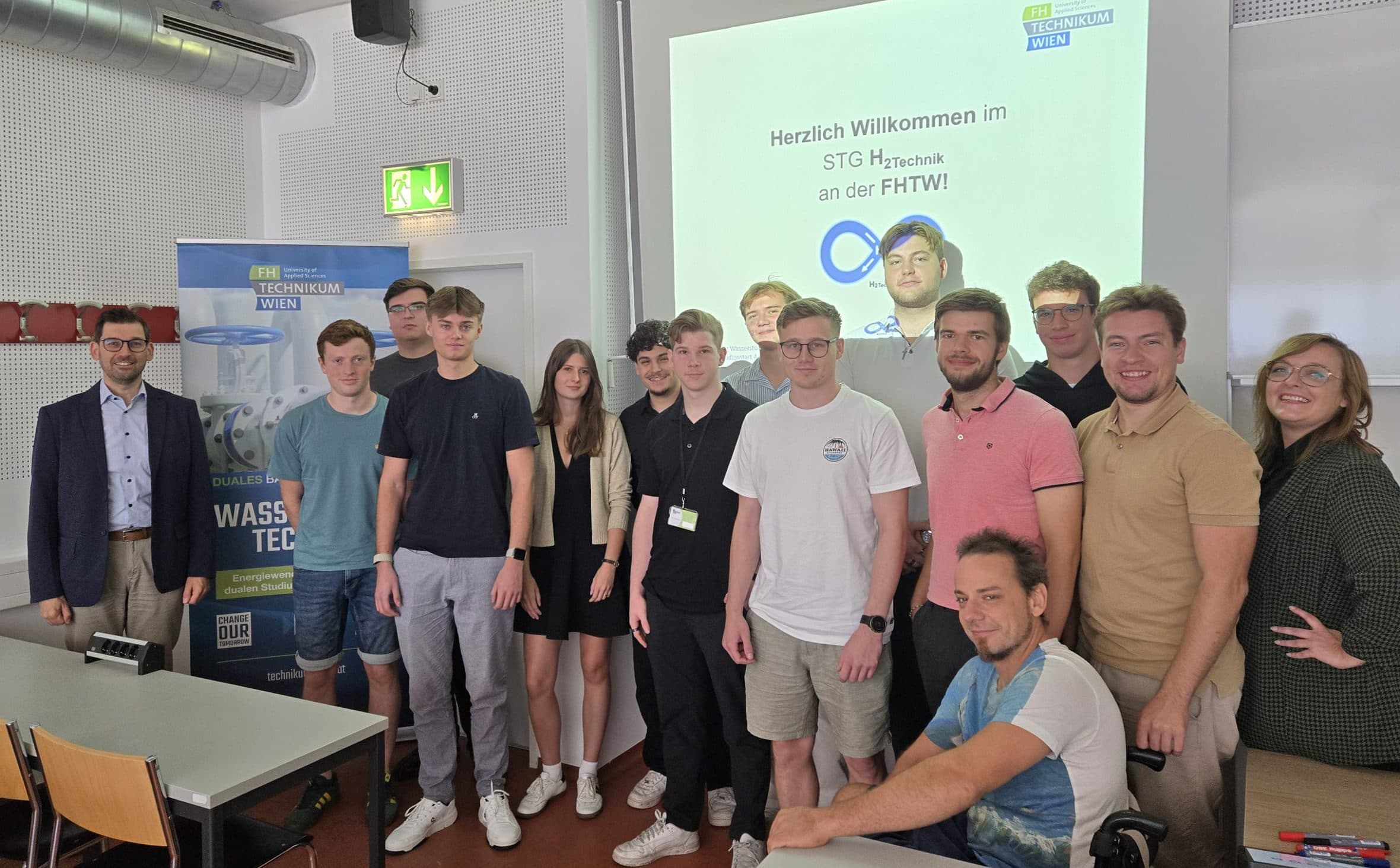 Eine Gruppe von dreizehn Personen, meist junge Erwachsene, steht in einem Klassenzimmer und lächelt. Auf einer Präsentationsfolie auf der Leinwand hinter ihnen steht "Herzlich Willkommen im STG Maschinenbau an der FH T." In dem Raum stehen Tische und Stühle.