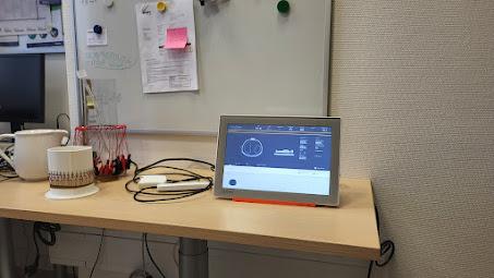 Auf einem hellen Holztisch steht ein Tablet, auf dem Daten angezeigt werden, neben einem Whiteboard, das mit Papieren und Magneten bedeckt ist. Eine Tasse, ein Behälter mit Stiften und verschiedene Kabel liegen ebenfalls auf dem Schreibtisch.