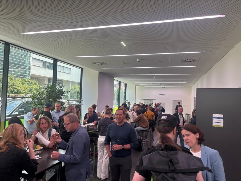 In einem modernen, sonnendurchfluteten Raum mit großen Fenstern und hohen Tischen trifft sich eine Gruppe von Menschen und isst, was auf ein zwangloses Networking oder ein Mittagessen hindeutet.