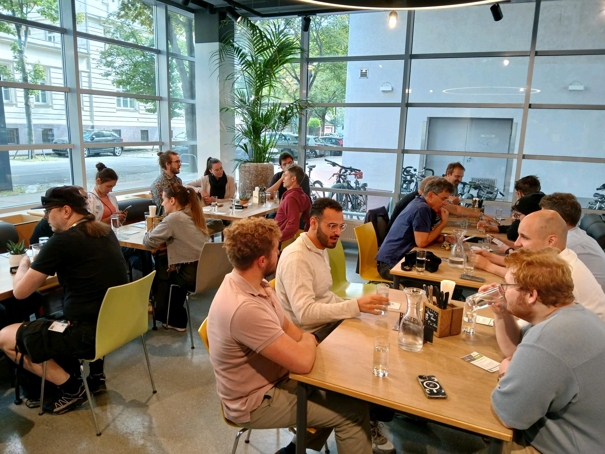 Eine Gruppe von Menschen sitzt an Tischen in einem modernen Café, unterhält sich und trinkt Wasser. Große Fenster zeigen Bäume, geparkte Fahrräder und Autos im Freien. Die Atmosphäre wirkt lebendig und gesellig.