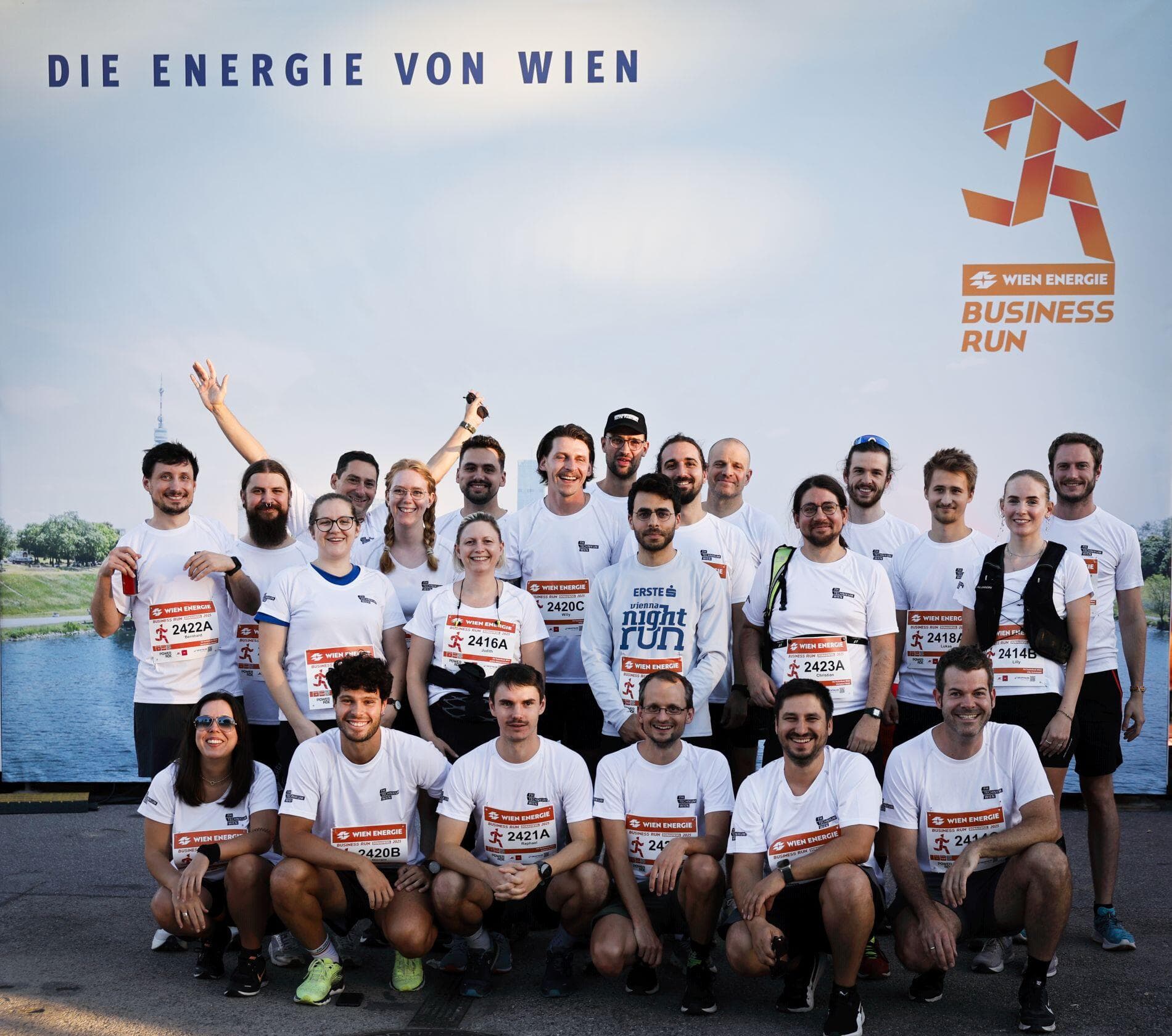 Eine Gruppe von Läufern in passenden weißen Hemden posiert und lächelt gemeinsam vor einem "Wien Energie Business Run"-Transparent in Wien. Die meisten tragen Startnummern und die Stimmung ist fröhlich und energiegeladen.