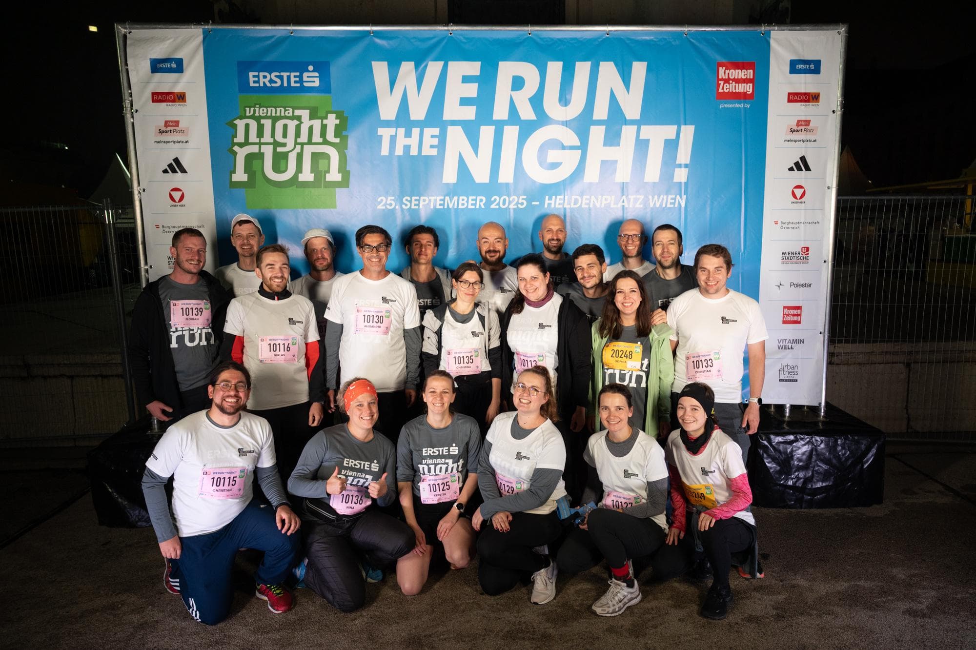 Eine Gruppe von Läufern posiert und lächelt vor einem nächtlichen Vienna Night Run-Banner. Die meisten tragen Startnummern und Sportkleidung und stehen und knien gemeinsam auf dem nassen Gehsteig.