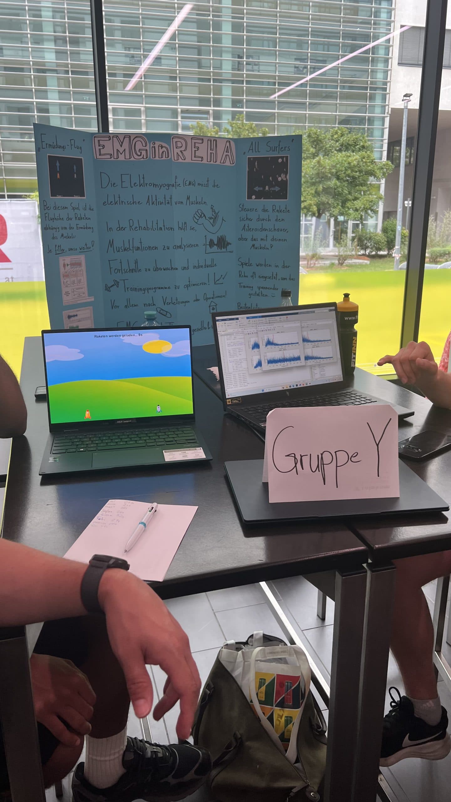 Auf einem Tisch stehen zwei Laptops, ein handgeschriebenes Schild der Gruppe Y, ein Notizbuch und eine Wasserflasche. Dahinter befindet sich eine blaue Präsentationstafel mit der Aufschrift EMG 4 REHA. Zwei Personen sind teilweise sichtbar, eine macht sich Notizen.