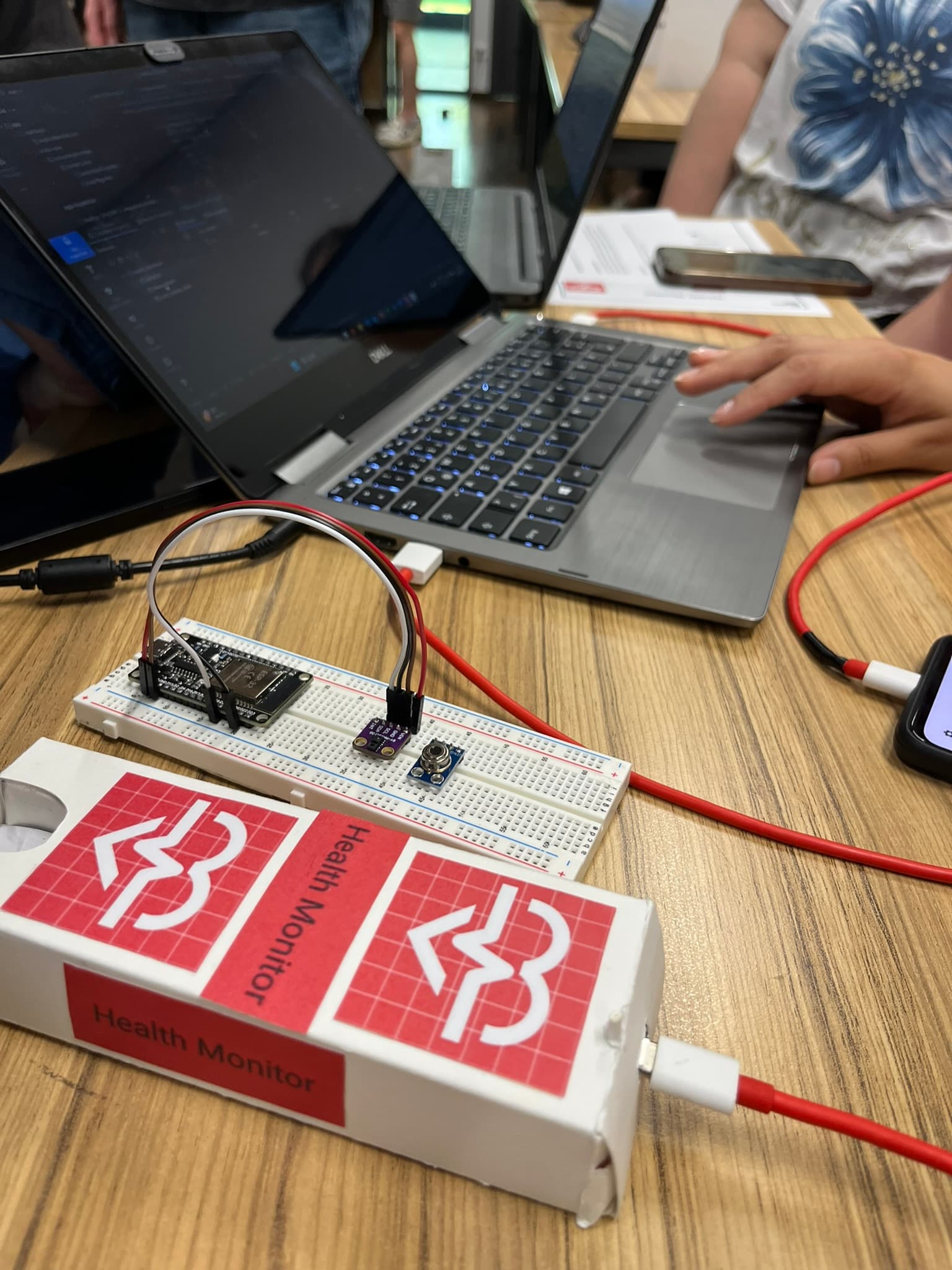 Nahaufnahme eines Gesundheitsüberwachungsgeräts, das an ein Breadboard mit elektronischen Komponenten angeschlossen ist, neben einem Laptop und einem Smartphone auf einem Holztisch. Eine Person tippt auf dem Laptop.