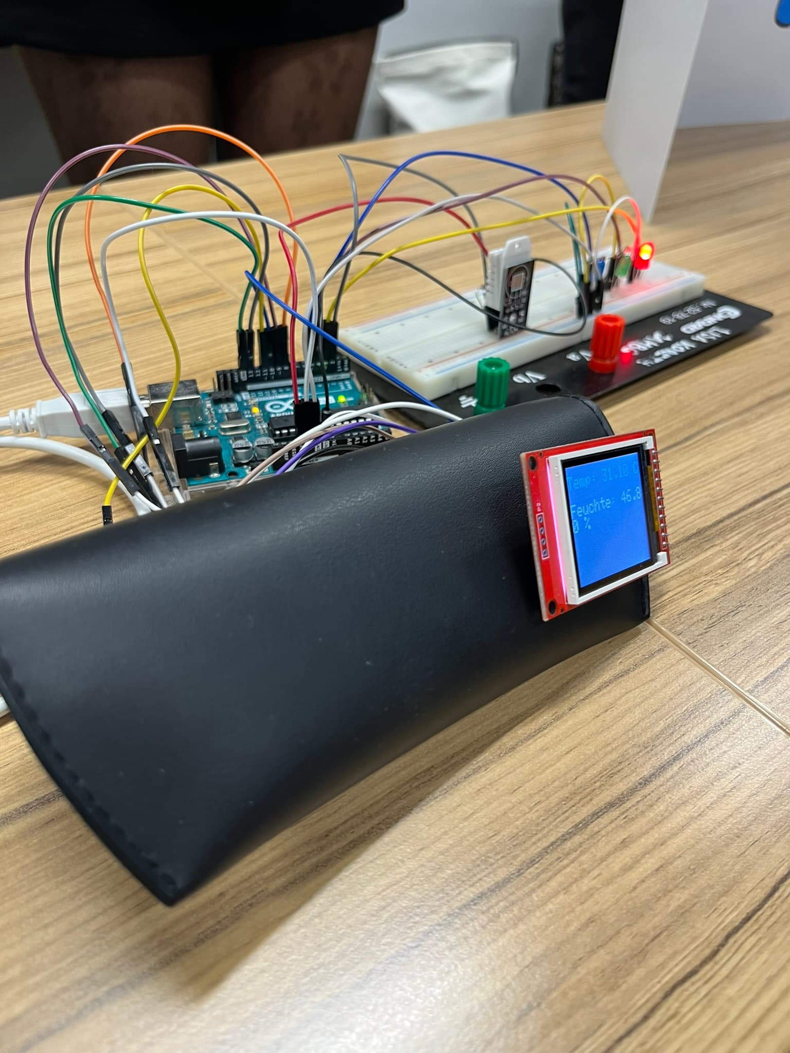 Ein elektronisches Projekt mit einem Arduino-Board, Drähten, einem Breadboard mit LEDs und einem kleinen Bildschirm, auf dem 'Graphite 46' angezeigt wird. Der Aufbau befindet sich auf einem Holztisch mit einem schwarzen Gehäuse unter dem Bildschirm.