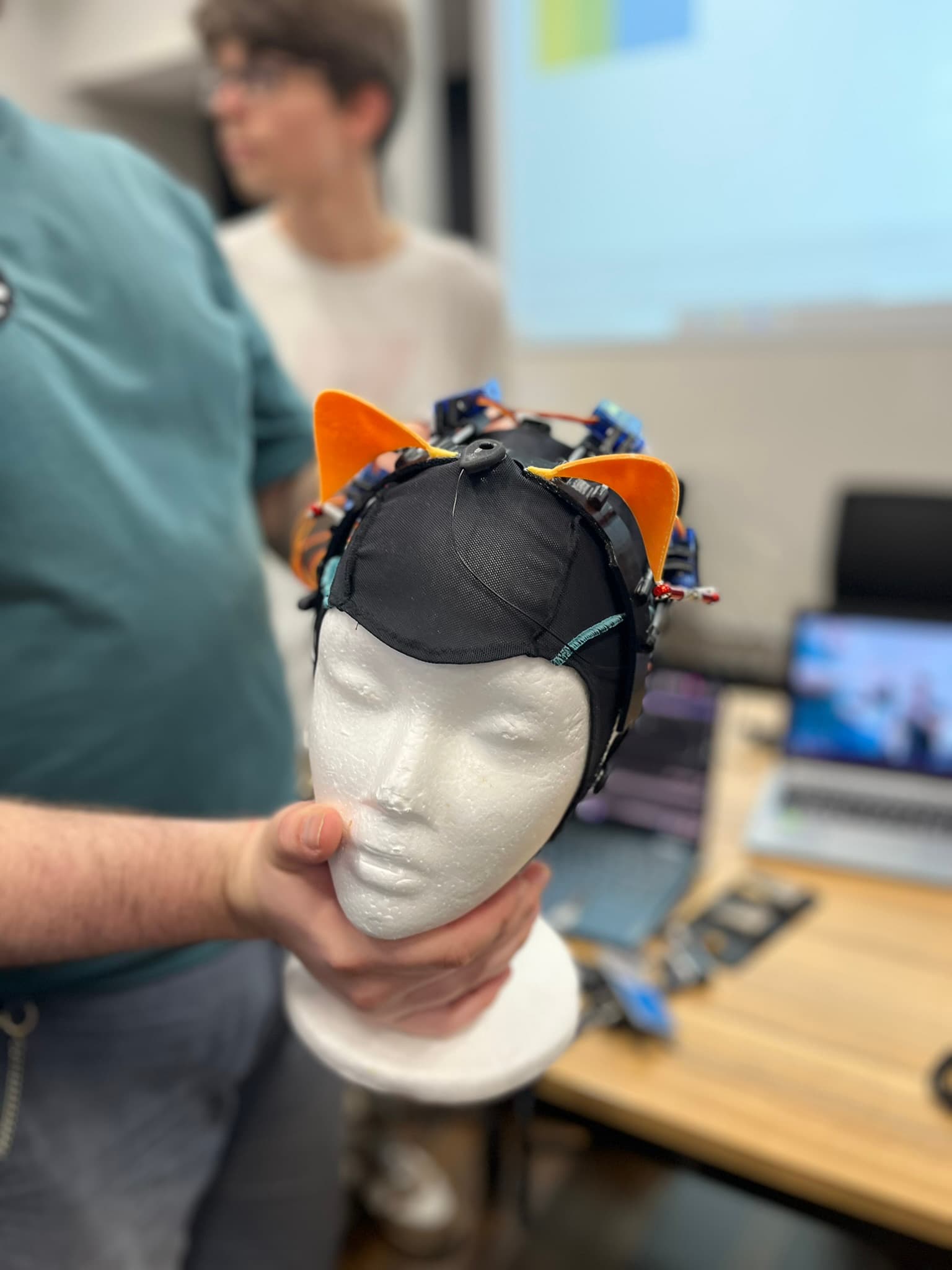Eine Person hält einen weißen Schaufensterpuppenkopf, der eine schwarze Mütze mit orangefarbenen Katzenohren und elektronischen Bauteilen trägt. Im Hintergrund sind eine verschwommene Person und elektronische Geräte zu sehen.