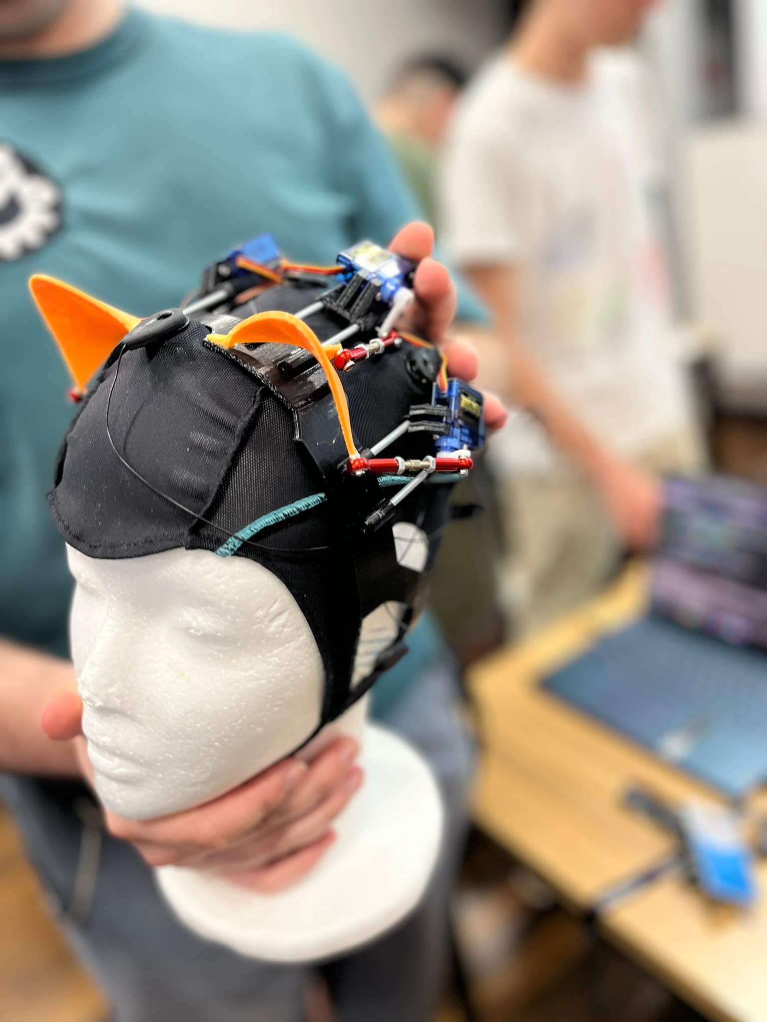 Eine Person hält eine Schaufensterpuppe, die eine schwarze Kappe trägt, die mit bunten Drähten, Sensoren und elektronischen Bauteilen bestückt ist; zwei Personen und ein Laptop sind unscharf im Hintergrund zu sehen.