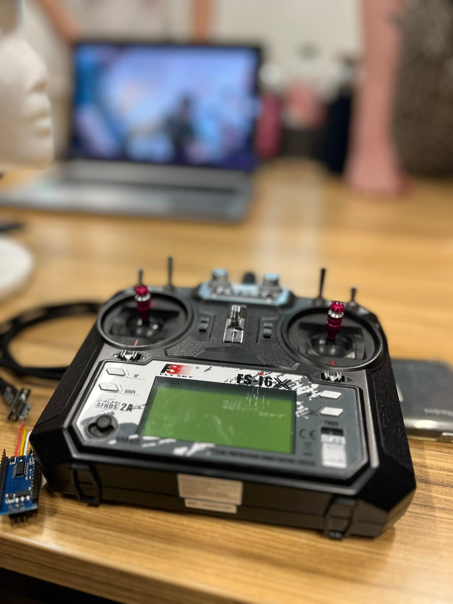 Eine schwarze Fernbedienung mit Joysticks und einem digitalen Bildschirm liegt auf einem Holztisch, im Hintergrund sind ein Laptop und unscharfe Objekte zu sehen.