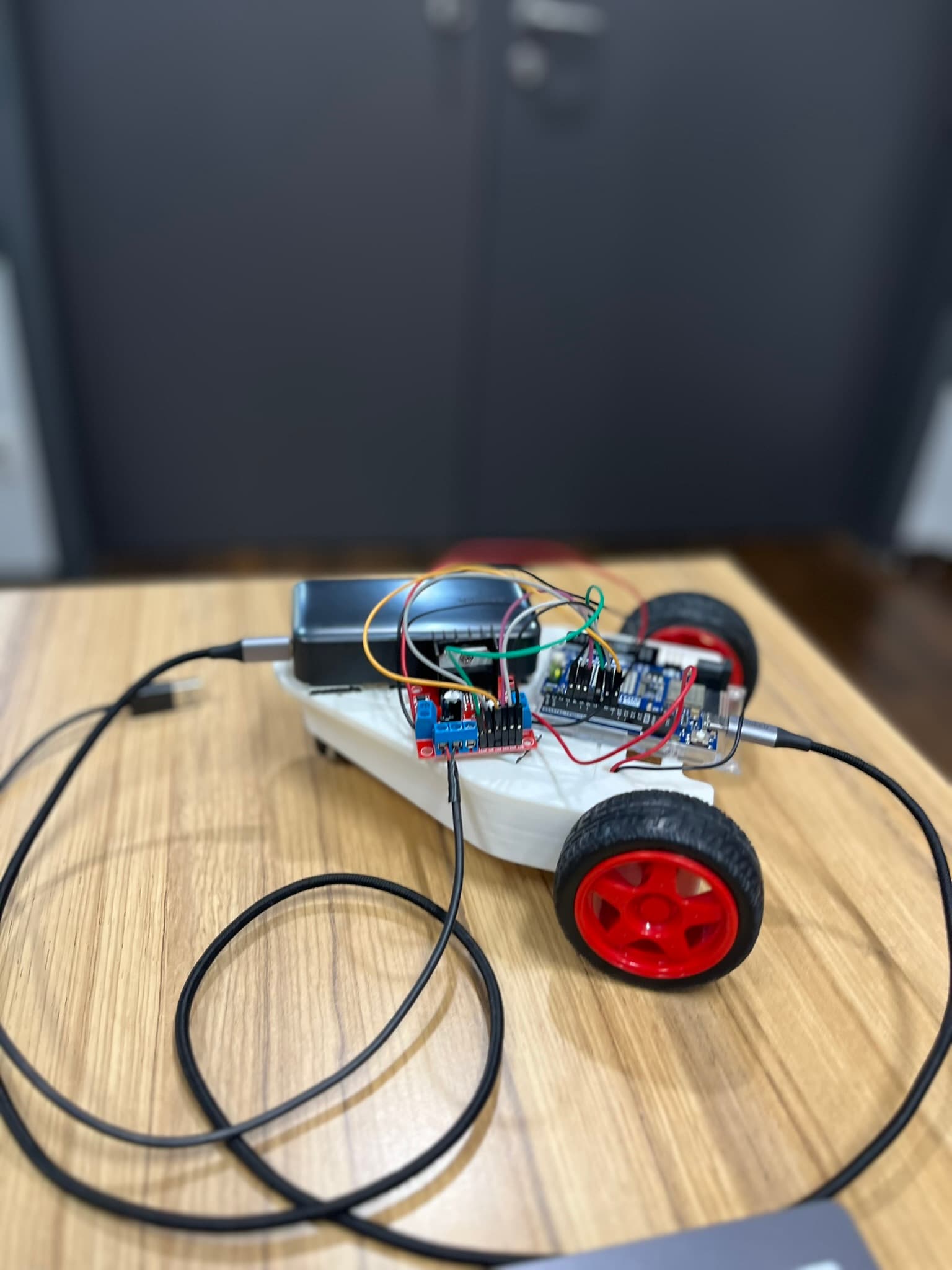 Ein kleines Roboterauto mit roten Rädern und freiliegenden elektronischen Bauteilen steht auf einem Holztisch und ist mit einem USB-Kabel verbunden. Der Hintergrund ist unscharf und zeigt eine graue Tür.