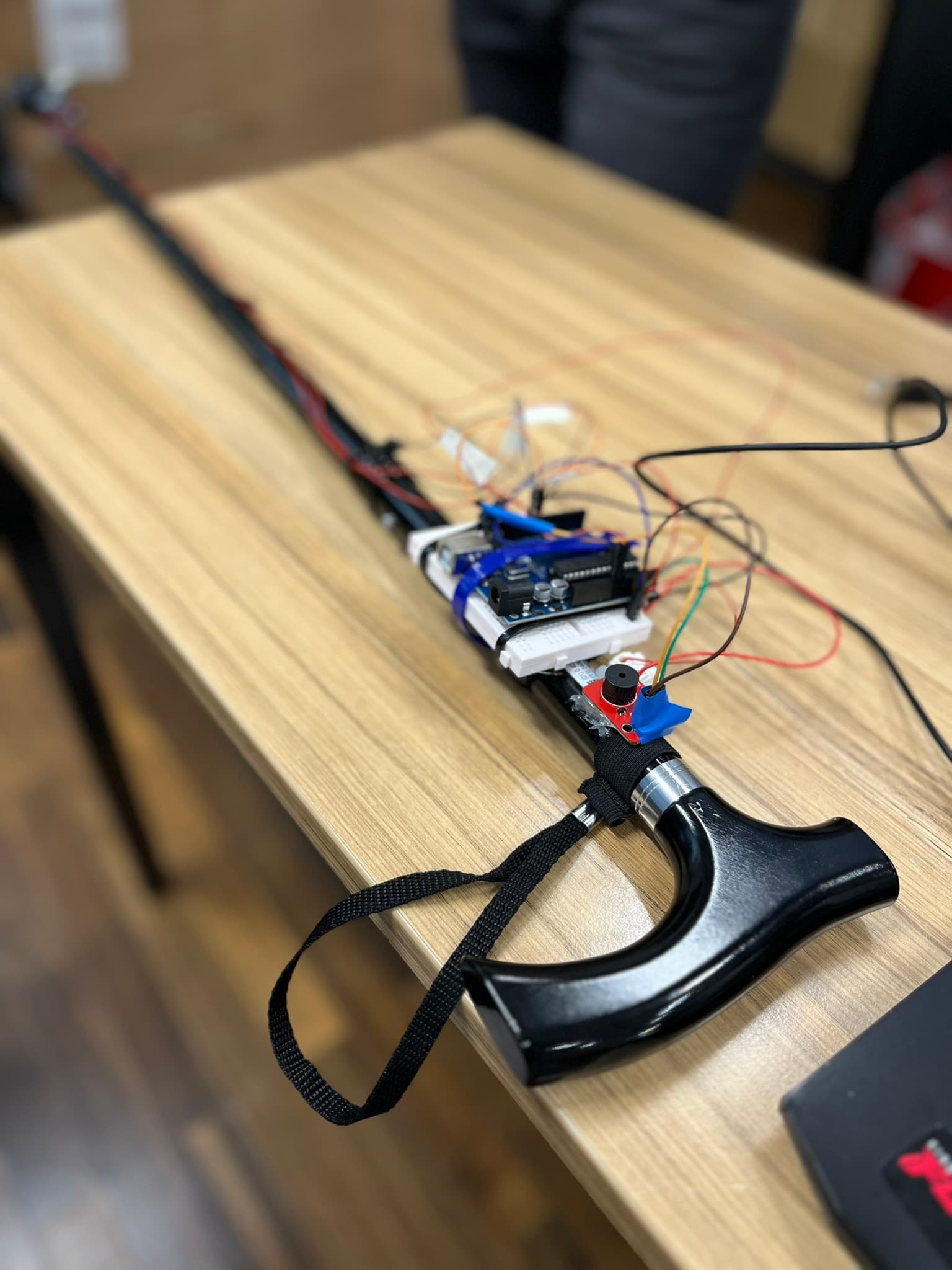 Ein schwarzer Gehstock mit elektronischen Bauteilen, Drähten und einer kleinen Platine in der Nähe des Griffs, der auf einem Holztisch liegt.