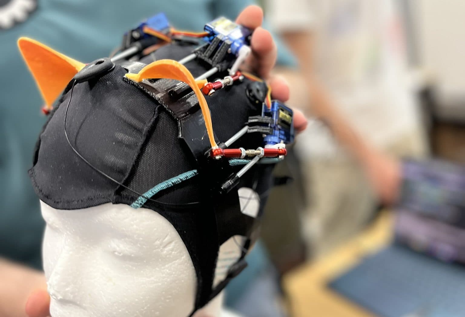 Eine Person hält eine weiße Schaufensterpuppe, die eine schwarze, mit elektronischen Bauteilen verkabelte Kappe und orangefarbene Katzenohren trägt, während im unscharfen Hintergrund zwei Personen und ein Laptop zu sehen sind.