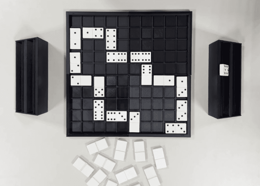Ein laufendes Dominospiel auf einem schwarzen Spielbrett mit Dominosteinen, die in verschiedene Richtungen gelegt werden. Zwei schwarze Ständer enthalten zusätzliche Dominosteine, und mehrere unbenutzte Dominosteine sind unter dem Brett verstreut.