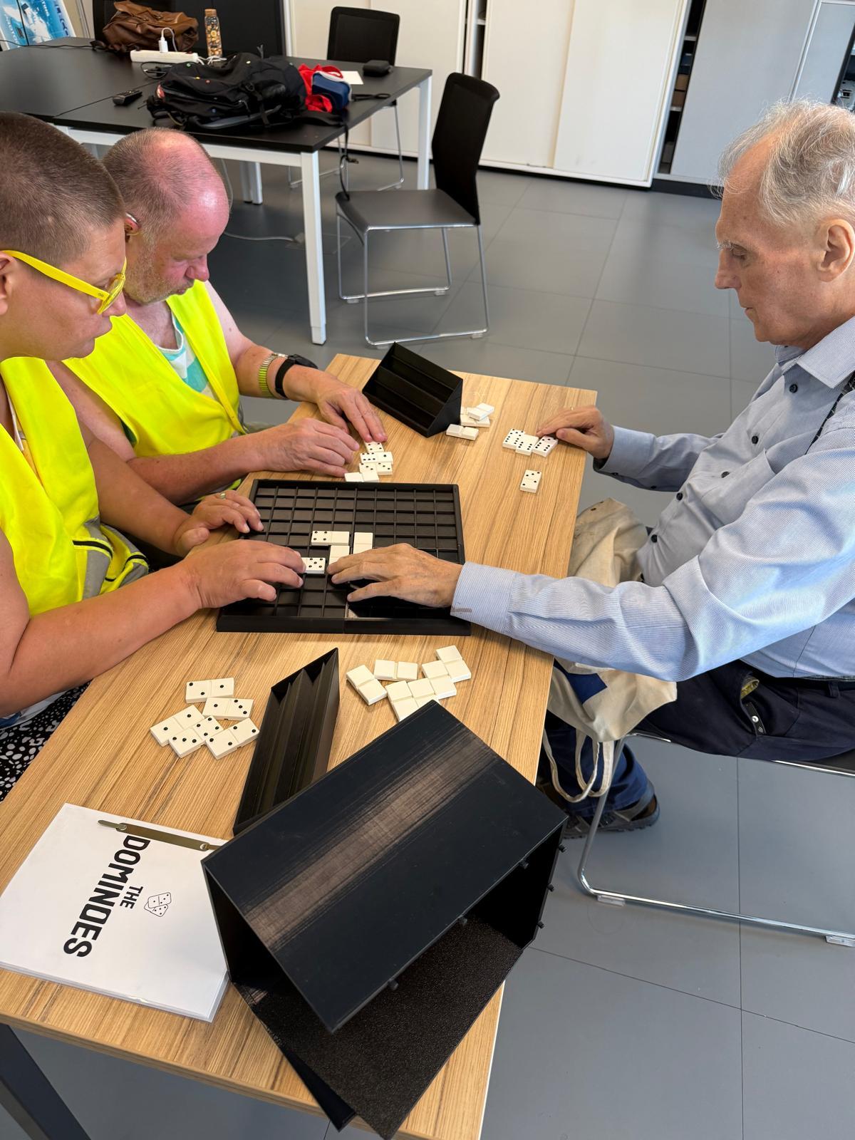 Drei Personen sitzen um einen Tisch und spielen eine taktile Version des Dominospiels. Zwei Spieler tragen gelbe Westen, und ein älterer Mann sitzt ihnen gegenüber. Auf dem Tisch liegen Dominosteine und Ständer, und ein Dominoheft ist sichtbar.