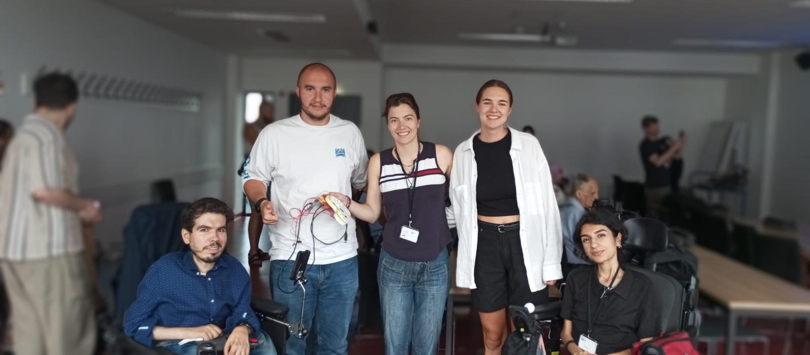 Fünf Personen, darunter zwei in Rollstühlen, posieren und lächeln gemeinsam in einem Klassenzimmer oder Konferenzraum. Andere Personen sind im Hintergrund zu sehen, und im Raum sind Tische und Stühle aufgestellt.