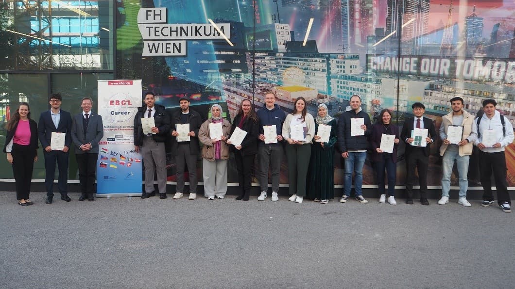 Eine Gruppe von Menschen steht in einer Reihe vor der FH Technikum Wien, hält Zertifikate in der Hand und lächelt in die Kamera. Auf der linken Seite ist ein Banner mit Fahnen und der Aufschrift "EBC*L Career" zu sehen. Im Hintergrund ist ein Wandgemälde mit einer Stadtlandschaft zu sehen.