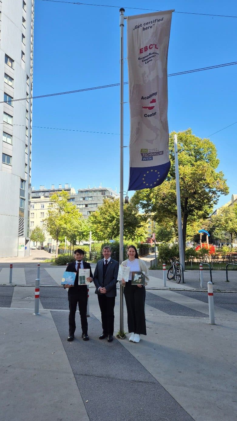 Drei Personen stehen im Freien vor einer großen Flagge mit Logos und Text. Zwei von ihnen halten Urkunden in der Hand. Im Hintergrund sind moderne Gebäude, Bäume und ein Spielplatz an einem sonnigen Tag zu sehen.