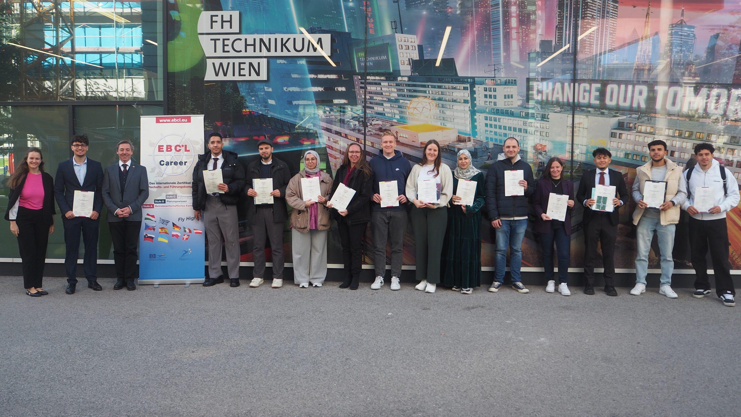 Eine Gruppe von Menschen steht in einer Reihe vor der FH Technikum Wien, hält Zertifikate in der Hand und lächelt. Sie stehen vor einem bunten Wandgemälde und einem Banner mit der Aufschrift 'EBCL Career'.