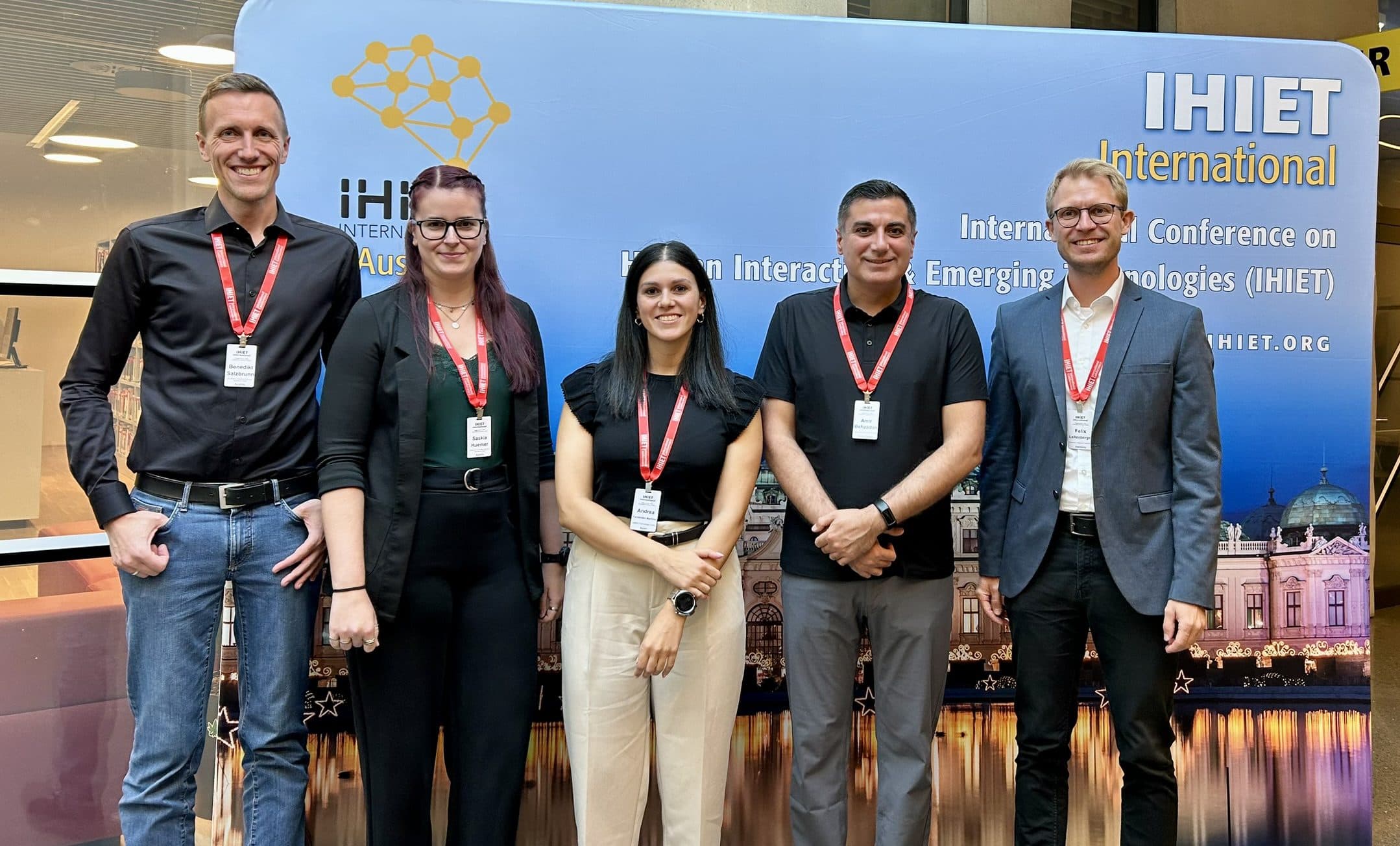 Fünf Personen stehen in einem Innenraum, lächeln in die Kamera, tragen Konferenzabzeichen und Geschäftskleidung. Hinter ihnen ist ein Banner für die internationale IHET-Konferenz zu sehen und eine nächtliche Stadtlandschaft im Hintergrund.
