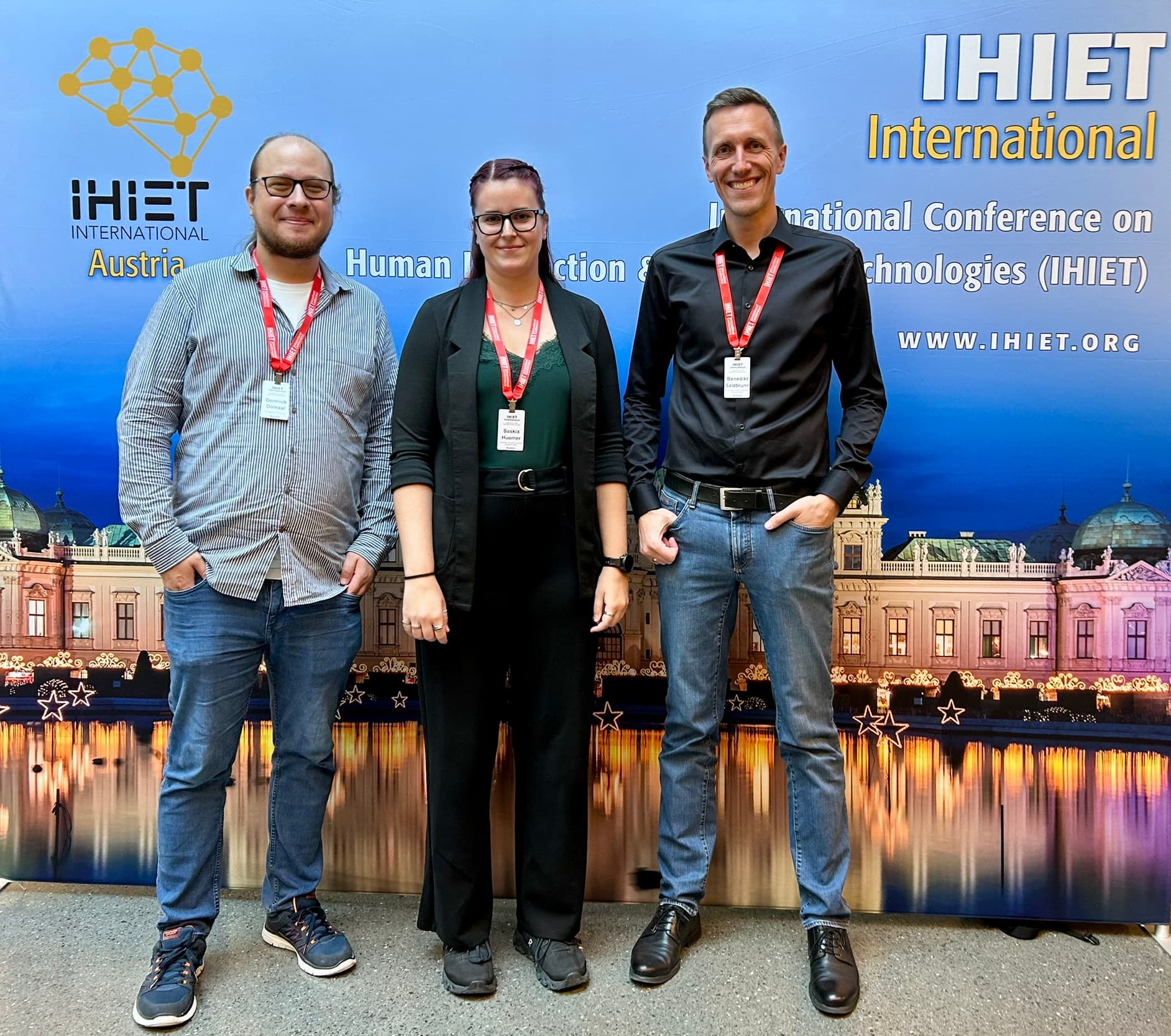 Drei Personen, die Konferenzabzeichen und rote Schlüsselbänder tragen, stehen lächelnd vor einem blauen IHIET International-Konferenzbanner, auf dem sich ein Gebäude mit Lichtern auf dem Wasser spiegelt.