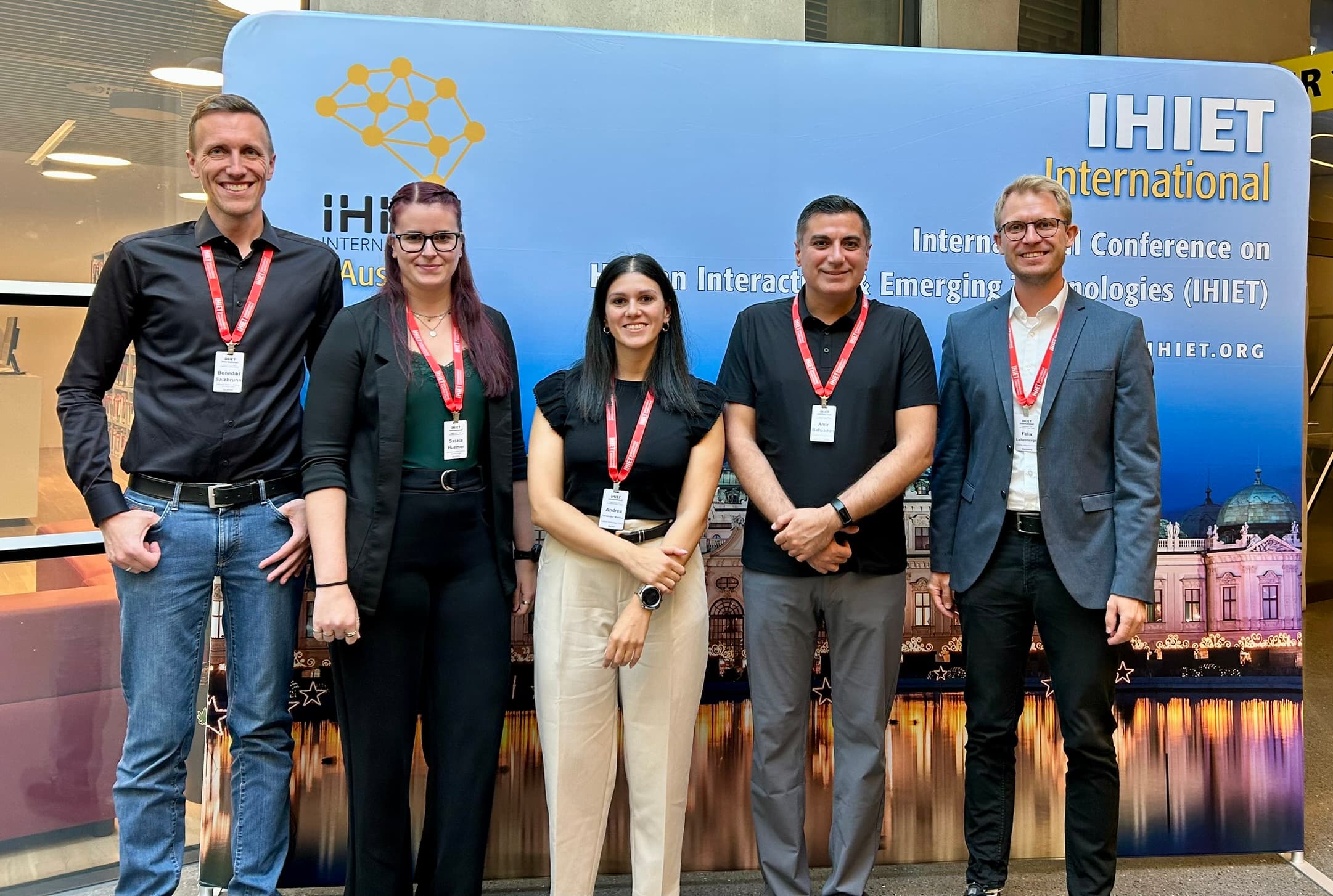 Fünf Personen stehen in einem Innenraum vor einem Banner der IHET International-Konferenz, alle tragen Namensschilder und legere Geschäftskleidung und lächeln in die Kamera. Im Hintergrund sind eine Stadtlandschaft und Veranstaltungsinformationen zu sehen.