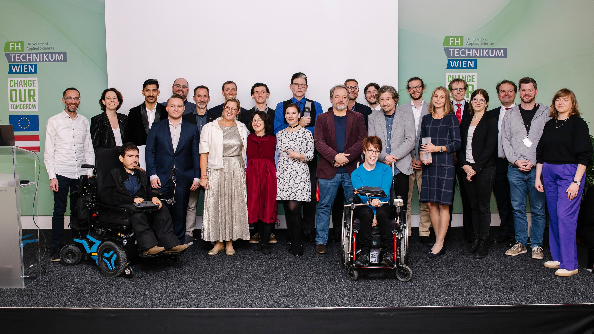 Eine Gruppe von etwa 25 Personen, darunter zwei Rollstuhlfahrer, posieren gemeinsam auf einer Bühne vor grünen Bannern mit den Logos der FH TECHNIKUM WIEN und Change Our Tomorrow. Alle lächeln in die Kamera.