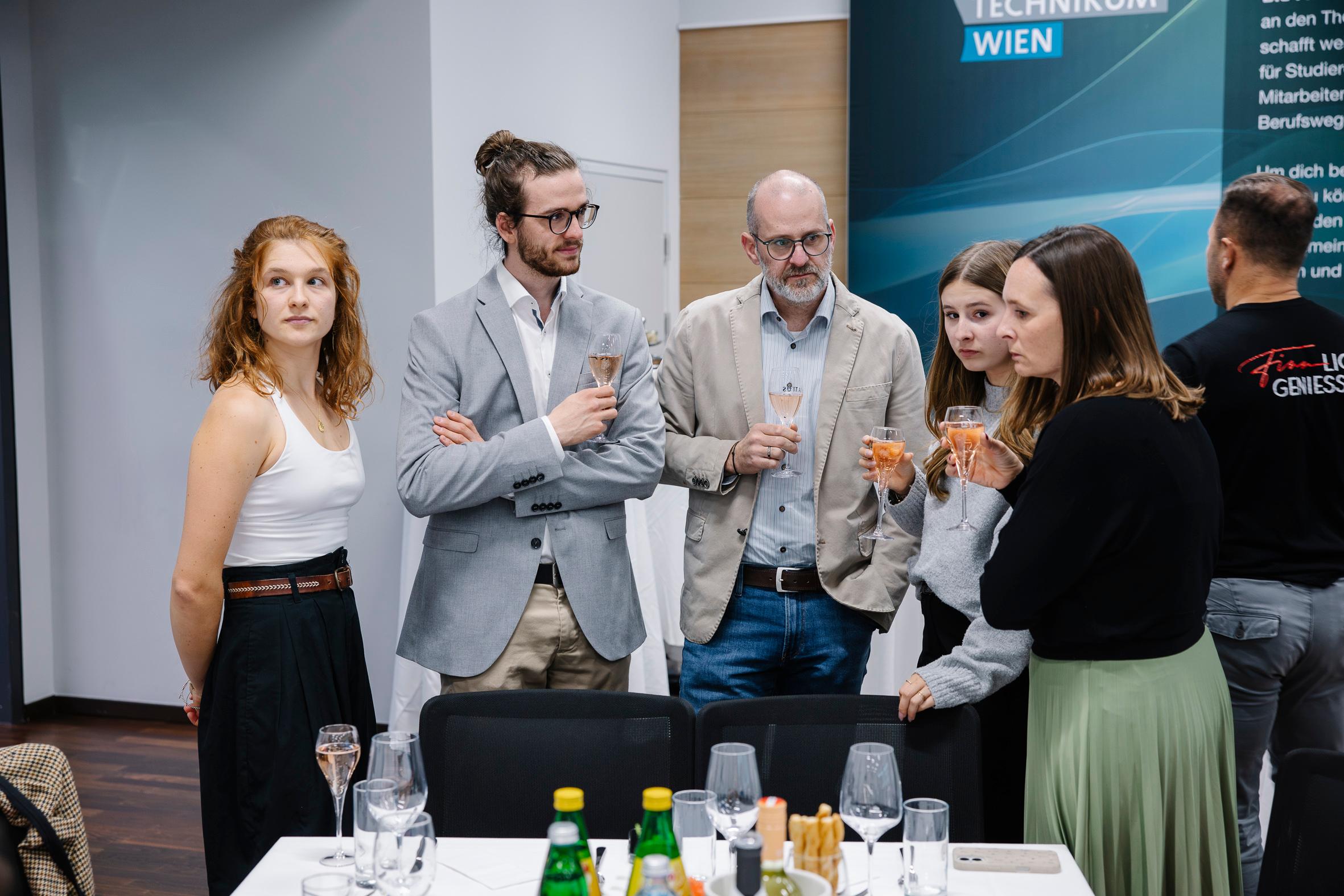 Sechs Personen stehen bei einer formellen Veranstaltung um einen Tisch herum, halten Getränke in der Hand und unterhalten sich. Die Gruppe ist in Business-Casual-Kleidung gekleidet. Flaschen, Gläser und Geschirr sind auf dem Tisch im Vordergrund zu sehen.