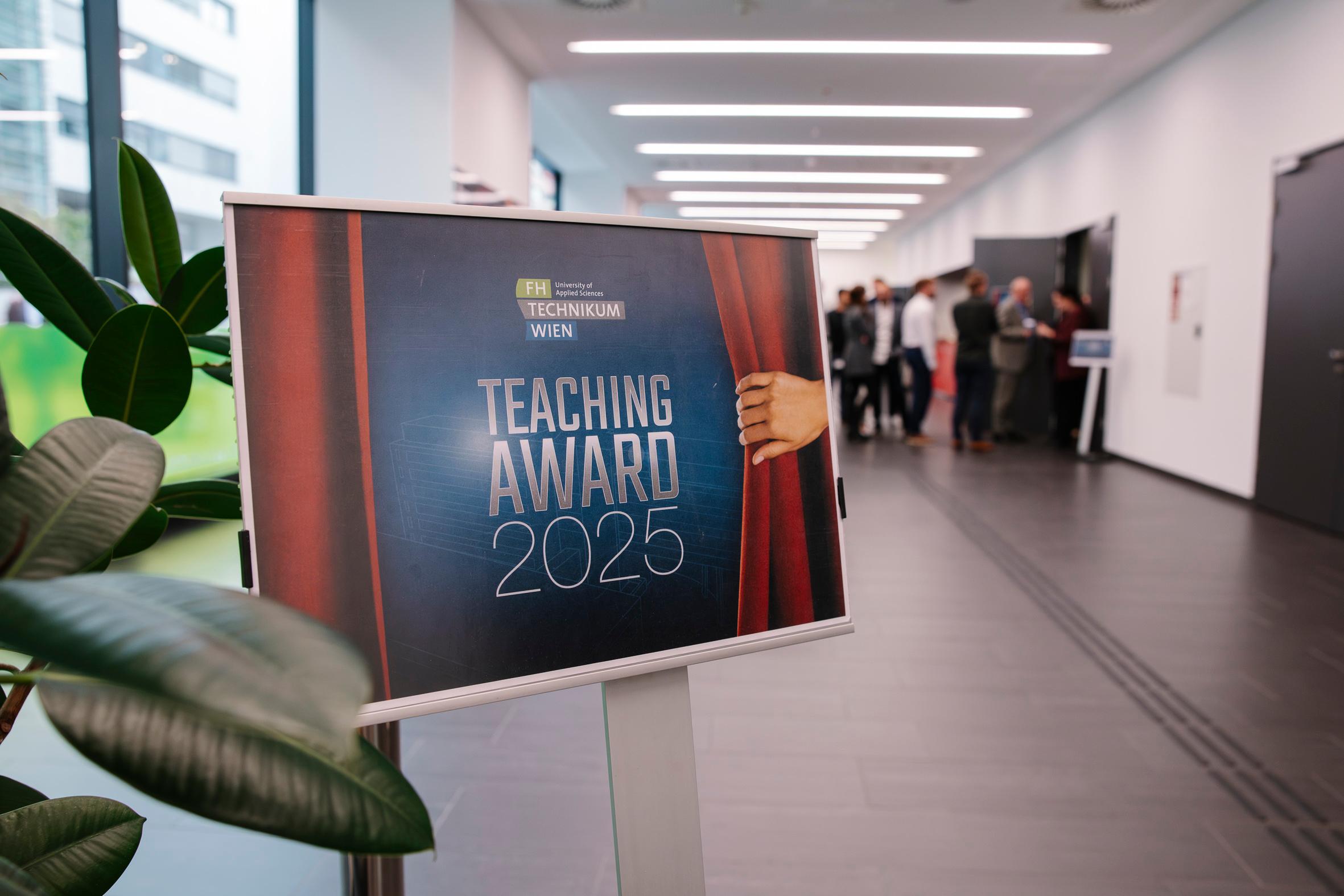Ein Schild mit der Aufschrift Teaching Award 2025 steht in einem Flur, in dem sich im Hintergrund Menschen versammeln und unterhalten. Im Vordergrund ist eine Pflanze zu sehen.