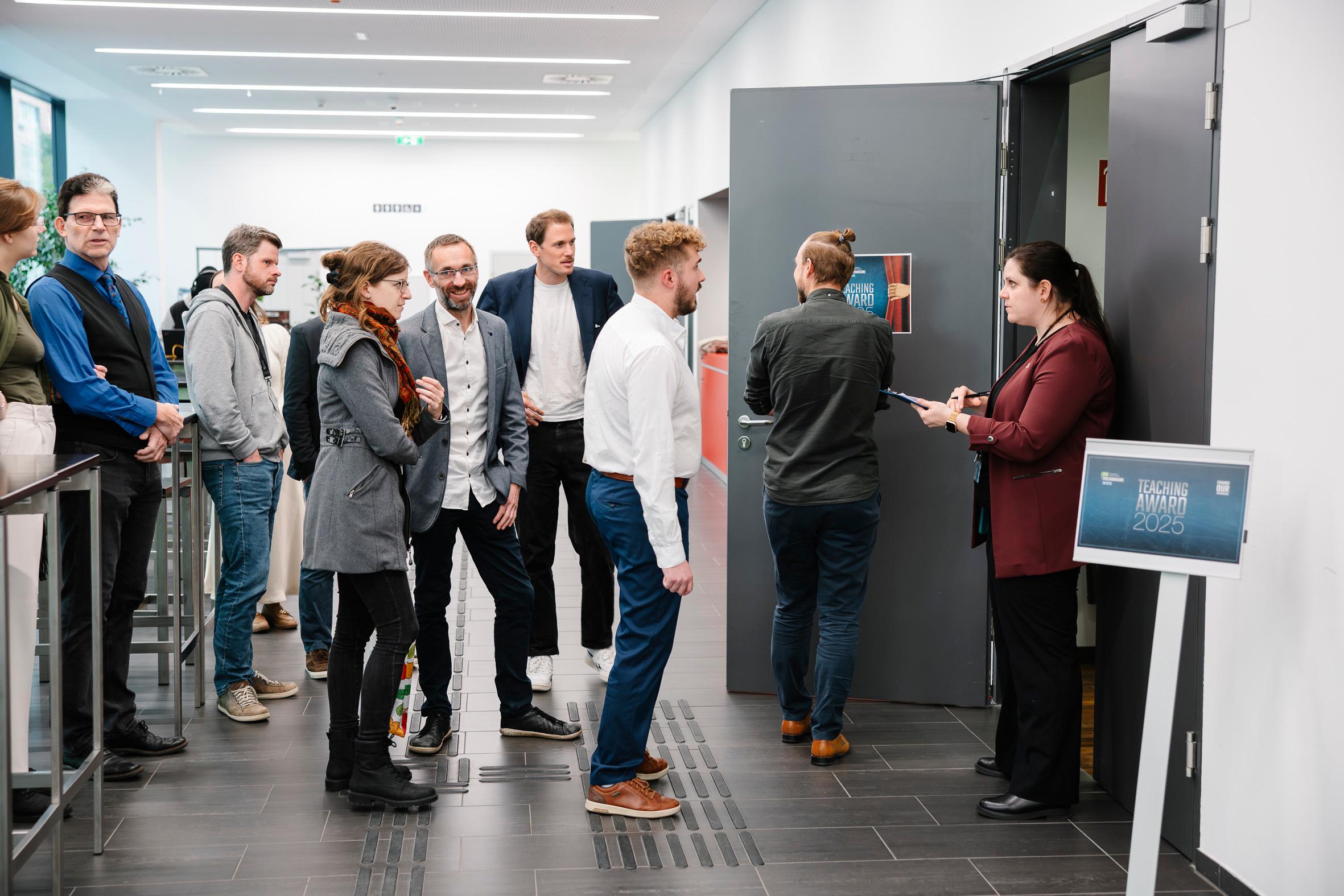 Eine Gruppe von Menschen steht in einer Schlange vor einem Raum, während eine Frau in einem kastanienbraunen Blazer die Eintrittskarten an der Tür kontrolliert. Auf einem Schild neben ihr steht 'EXAM AWARD 2025'. Der Schauplatz scheint ein moderner Flur zu sein.