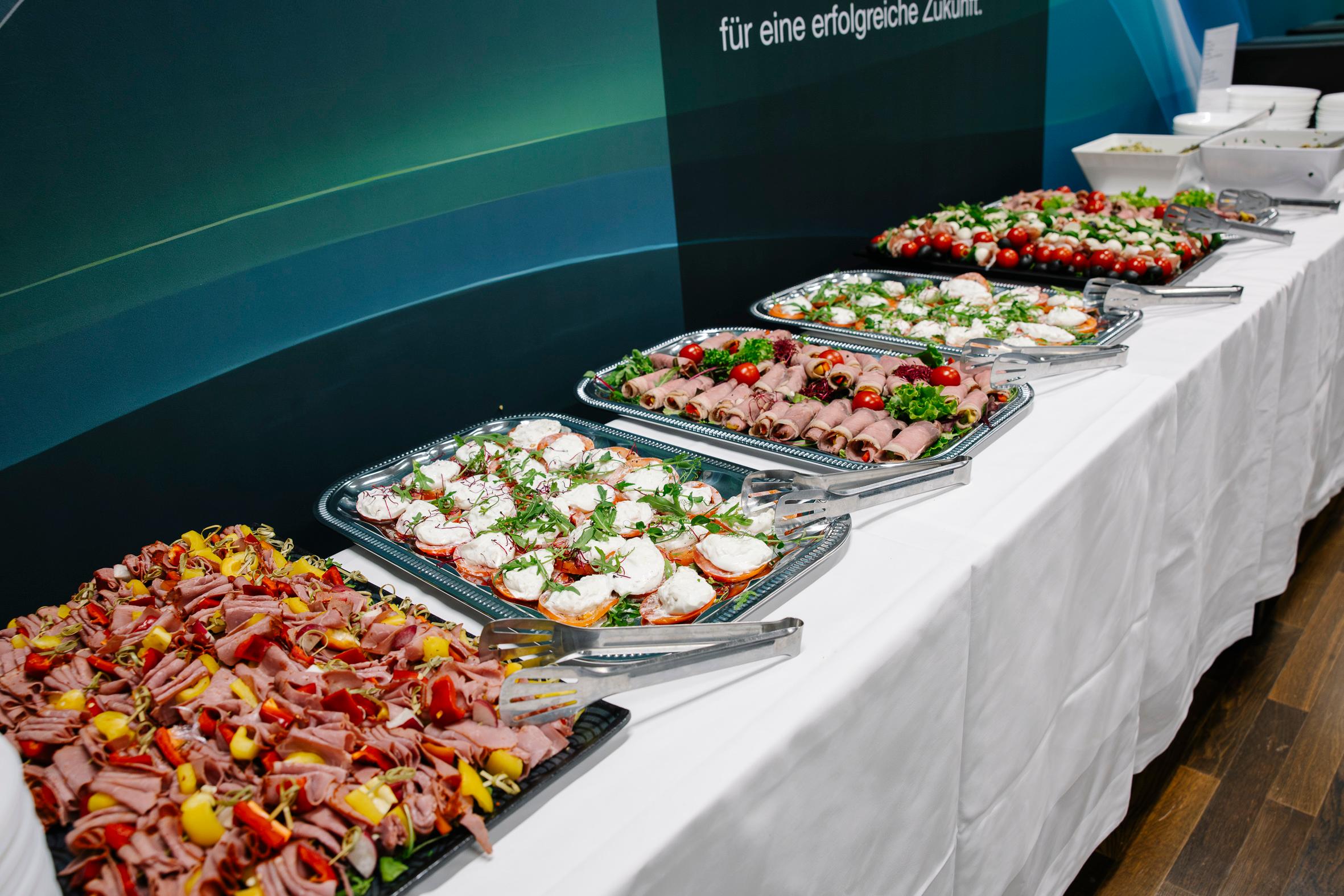 Ein Buffet mit verschiedenen Aufschnitt-, Fleisch-, Sandwich- und Gemüseplatten auf einem weißen Tischtuch mit Servierzangen und Salatschüsseln im Hintergrund.