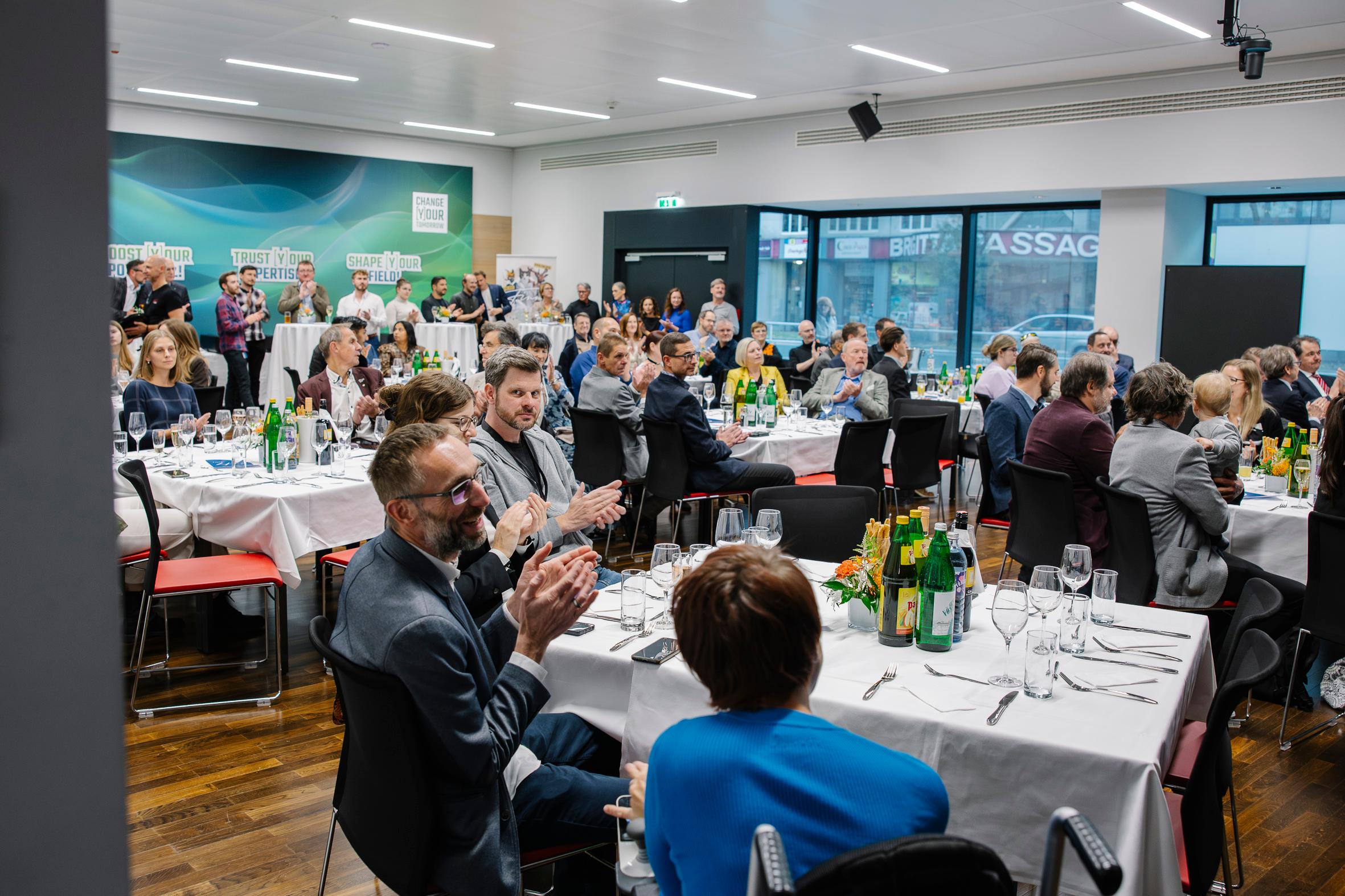 Eine Gruppe von Menschen sitzt an runden, mit weißen Tischtüchern bedeckten Tischen, klatscht und hört einem Redner bei einer formellen Veranstaltung im Haus zu. Flaschen und Gläser sind auf den Tischen angeordnet, andere stehen im Hintergrund.