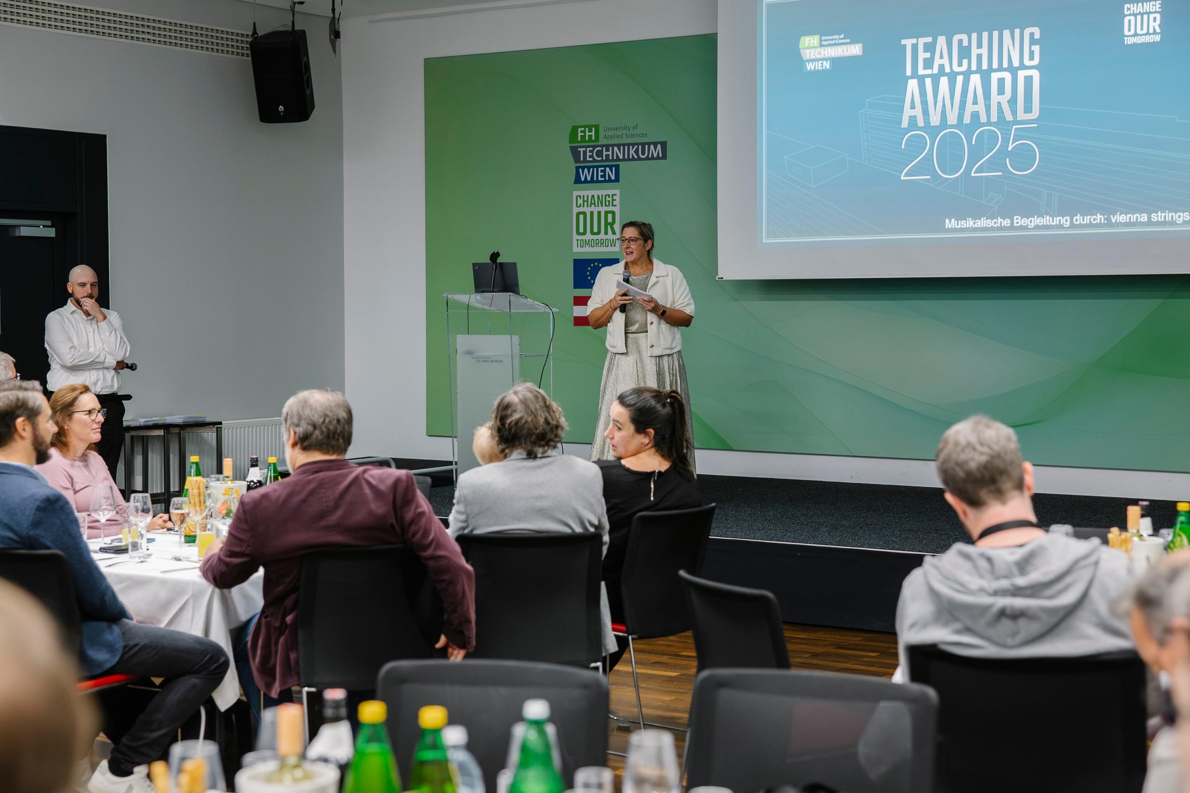 Eine Frau steht auf der Bühne und spricht an einem Podium während der Veranstaltung zum FH Technikum Wien Teaching Award 2025, während die Gäste an Tischen mit Getränken und Speisen sitzen und zuhören. Hinter ihr ist eine Präsentationsfolie zu sehen.