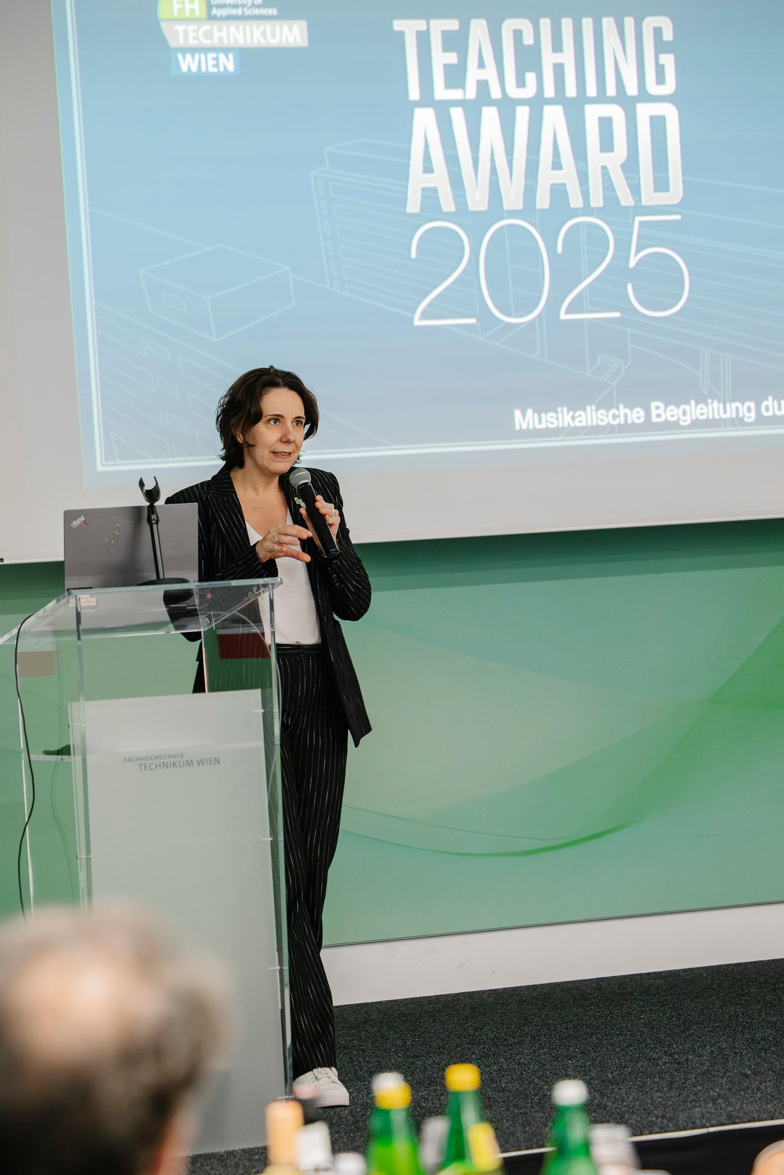 Eine Frau in einem schwarzen Nadelstreifenanzug spricht während der Veranstaltung zum FH Technikum Wien Teaching Award 2025 in ein Mikrofon am Podium. Hinter ihr wird eine Präsentationsfolie in einen modernen Konferenzraum projiziert.