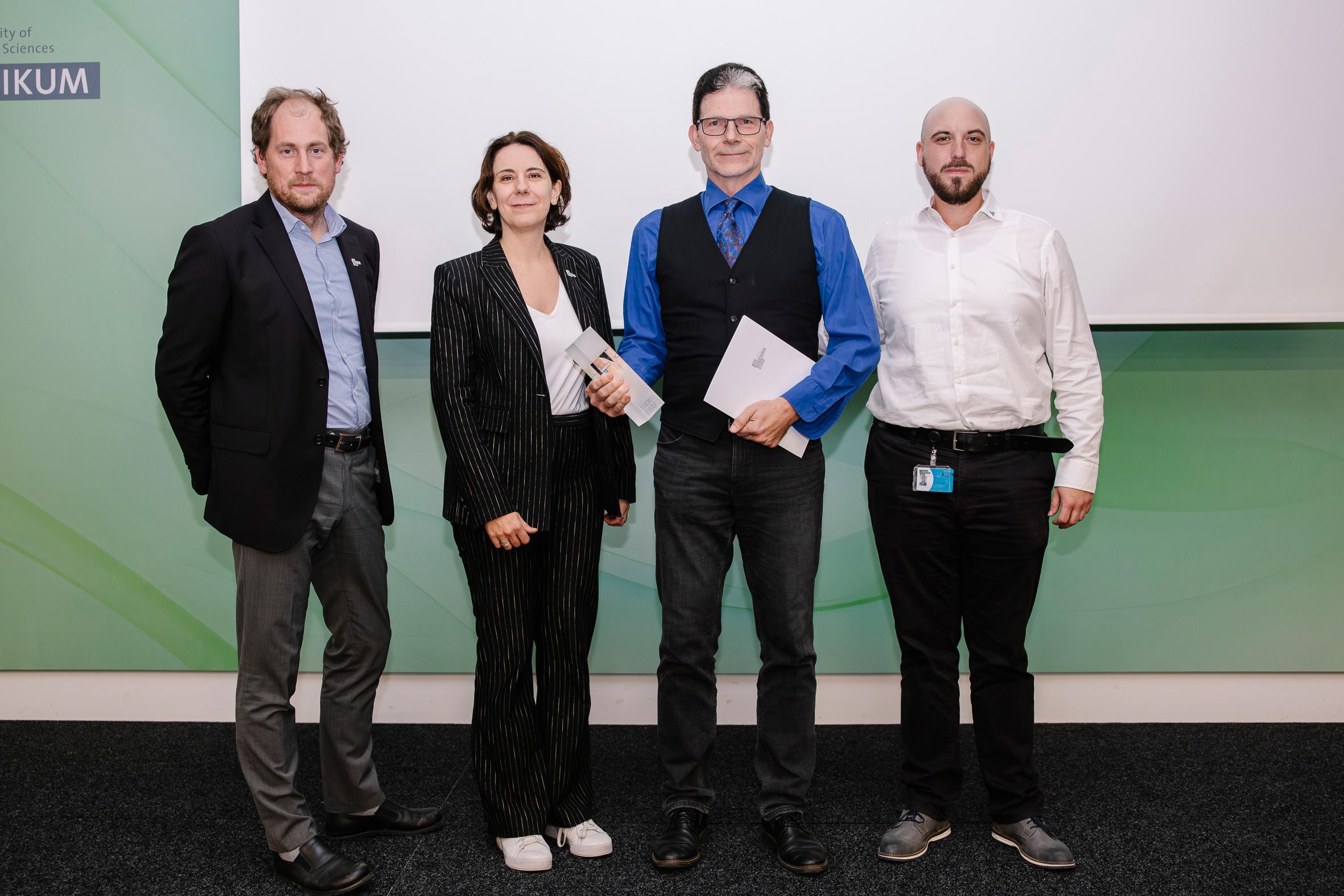Vier Personen in Geschäftskleidung stehen nebeneinander, zwei halten Umschläge in der Hand, vor einer grün-weißen Wand mit einem teilweise sichtbaren Logo auf der linken Seite. Sie posieren für ein Gruppenfoto in einem Innenraum.