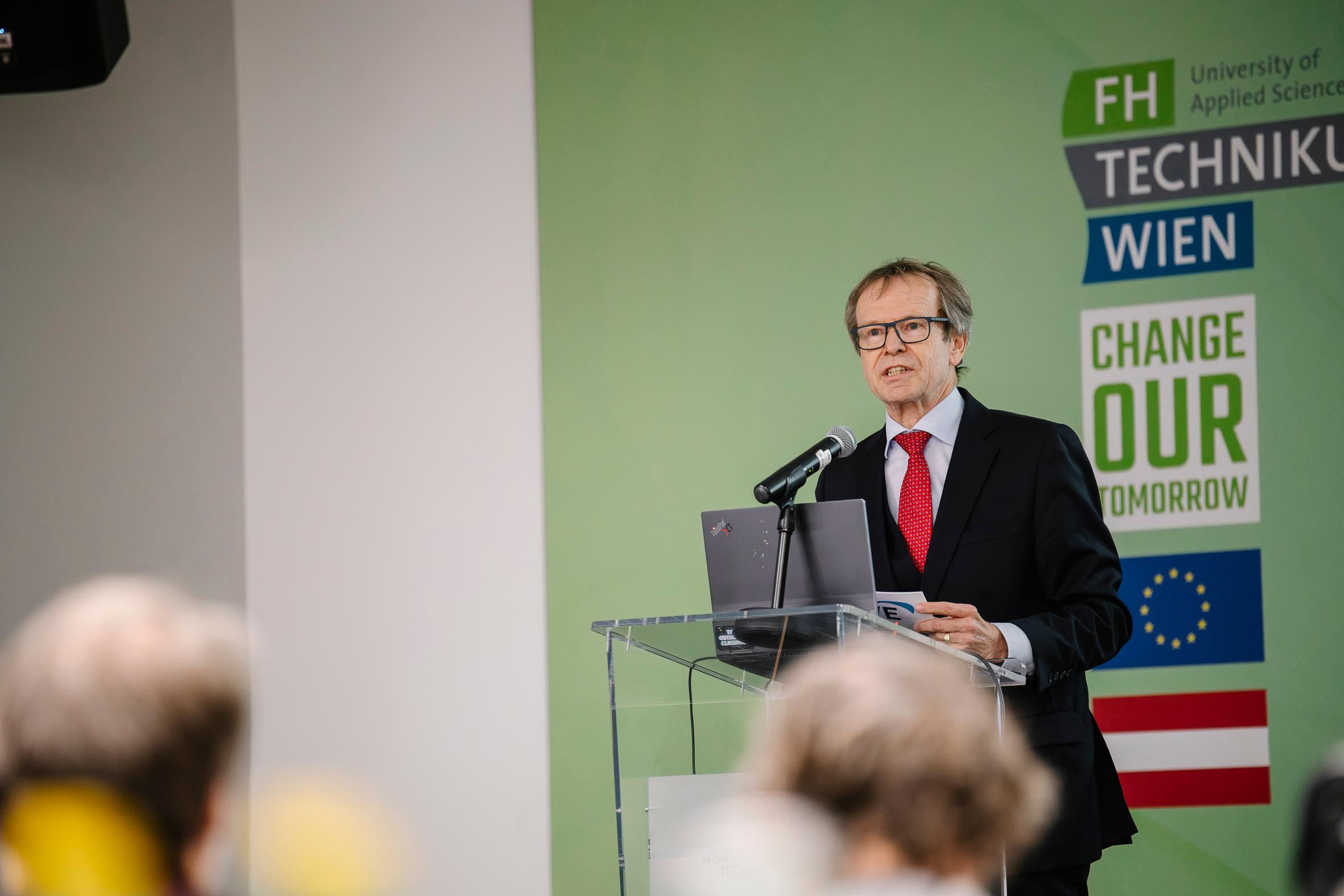 Ein Mann im Anzug und mit roter Krawatte spricht an einem Podium mit Laptop und Mikrofon. Hinter ihm befindet sich eine grüne Wand mit Logos und der Aufschrift 'FH Technikum Wien' und 'Change Our Tomorrow' sowie den Flaggen der EU und Österreichs.