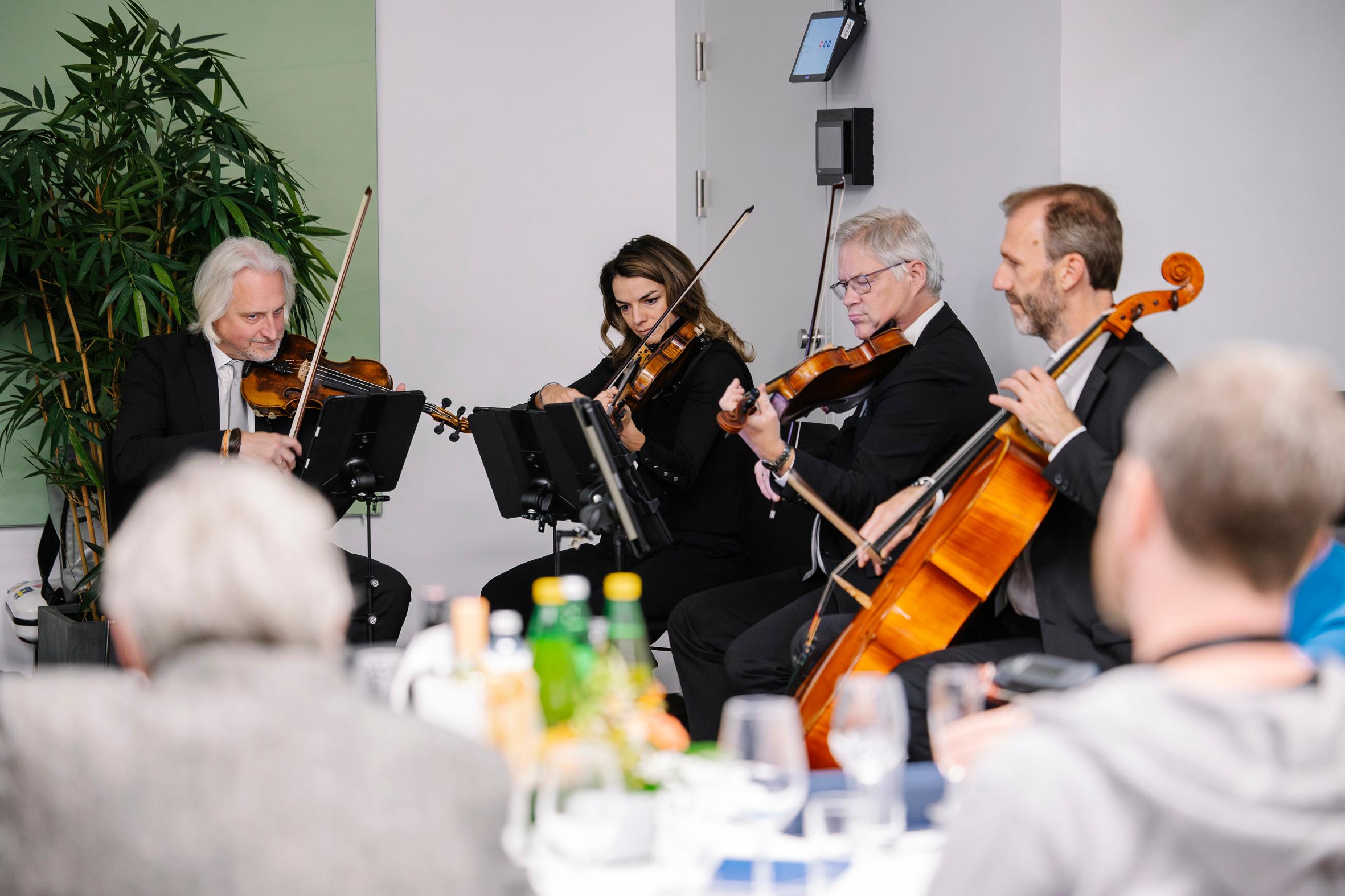 Ein Streichquartett tritt bei einer Indoor-Veranstaltung auf; drei Männer und eine Frau spielen Geigen und ein Cello, in formeller Kleidung sitzend, mit einem Publikum und einem gedeckten Tisch im Vordergrund.