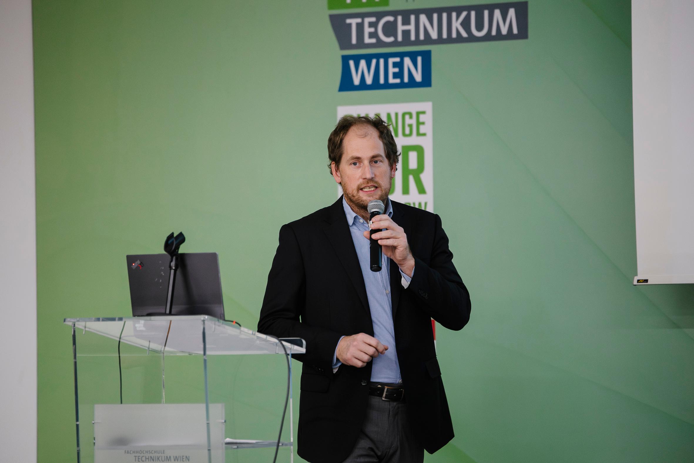 Ein Mann im Anzug, der ein Mikrofon hält, spricht an einem Podium mit einem Laptop. Im Hintergrund ist eine grüne Wand mit dem Schriftzug TECHNIKUM WIEN und anderem Text teilweise sichtbar.