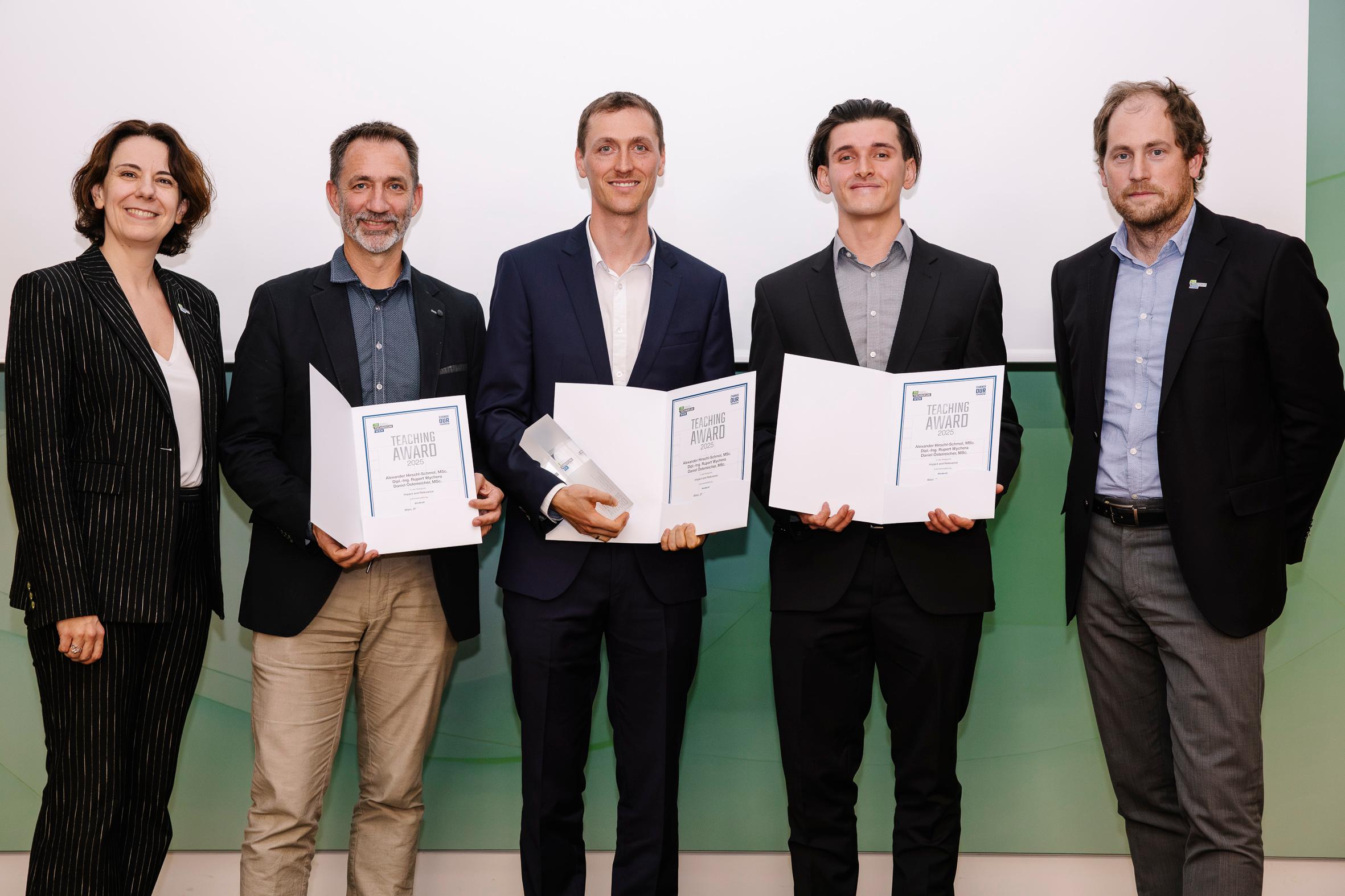 Fünf Personen stehen in einer Reihe, drei halten Urkunden und eine eine Glastrophäe in der Hand. Alle sind in Geschäftskleidung gekleidet, lächeln und posieren für ein Gruppenfoto vor einem hellen Hintergrund.