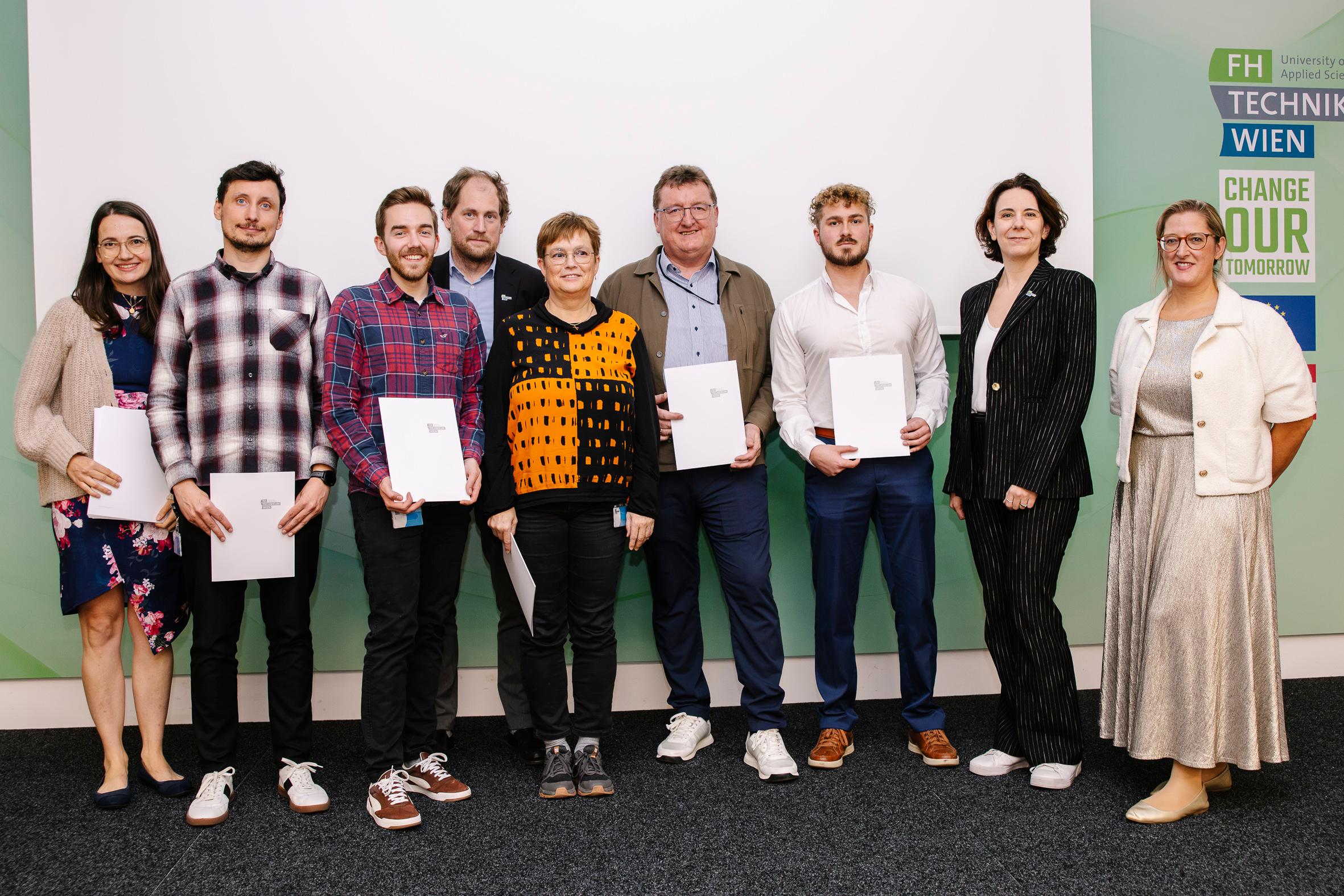 Acht Personen stehen in einem Innenraum in einer Reihe, einige mit Zertifikaten in der Hand, vor einem weißen Bildschirm und einer Wand mit den Logos der FH Technikum Wien und Change Your Tomorrow. Alle sind in Business- oder Smart-Casual-Kleidung gekleidet.