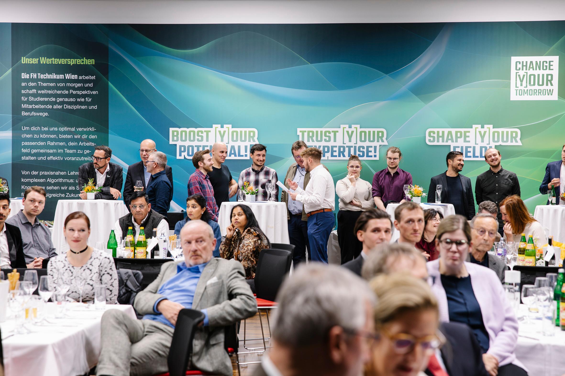 Eine Gruppe von Personen, sowohl sitzend als auch stehend, nimmt an einer formellen Veranstaltung in einem modernen Veranstaltungsort teil. Hinter ihnen sind grüne Wandgrafiken mit Sätzen zu sehen: BOOST YOUR POTENTIAL, TRUST YOUR EXPERTISE! und SHAPE YOUR EXPERIENCE!.