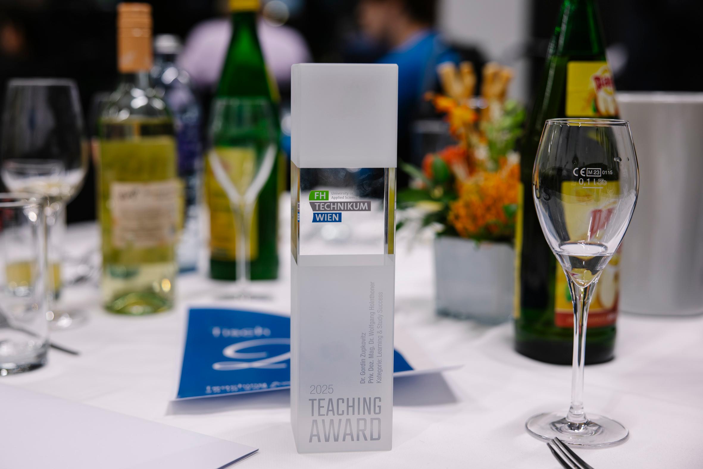 Eine Trophäe des Lehrpreises 2025 der FH Technikum Wien steht auf einem Tisch mit Weinflaschen, Gläsern, Blumen und blauen Broschüren, während die Menschen im Hintergrund verschwimmen.