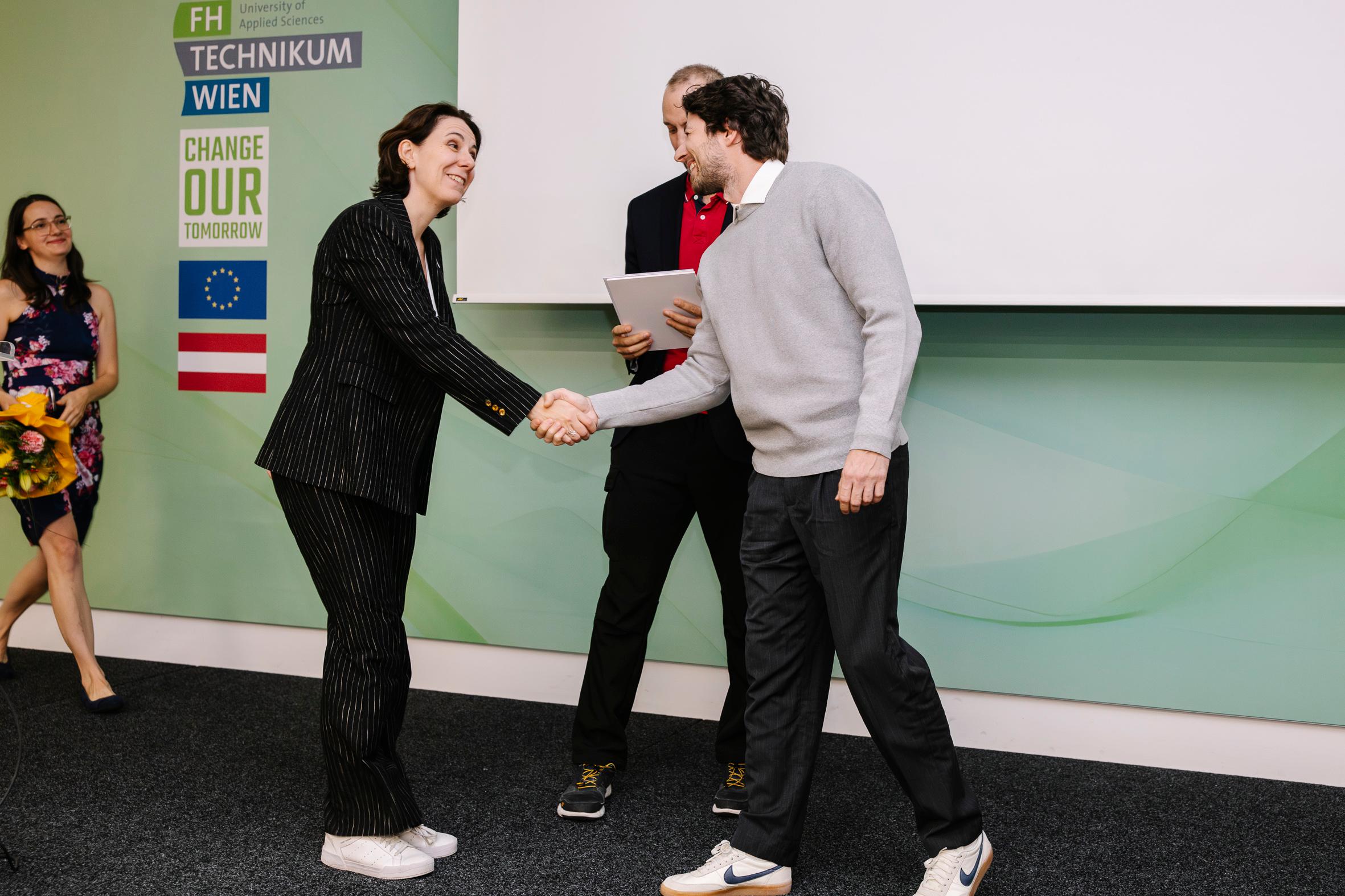 Eine Frau in einem Nadelstreifenanzug schüttelt während einer Veranstaltung einem Mann in einem grauen Pullover die Hand. Ein anderer Mann steht hinter ihnen und hält einen Ordner. Im Hintergrund ist eine Frau mit Blumen zu sehen.