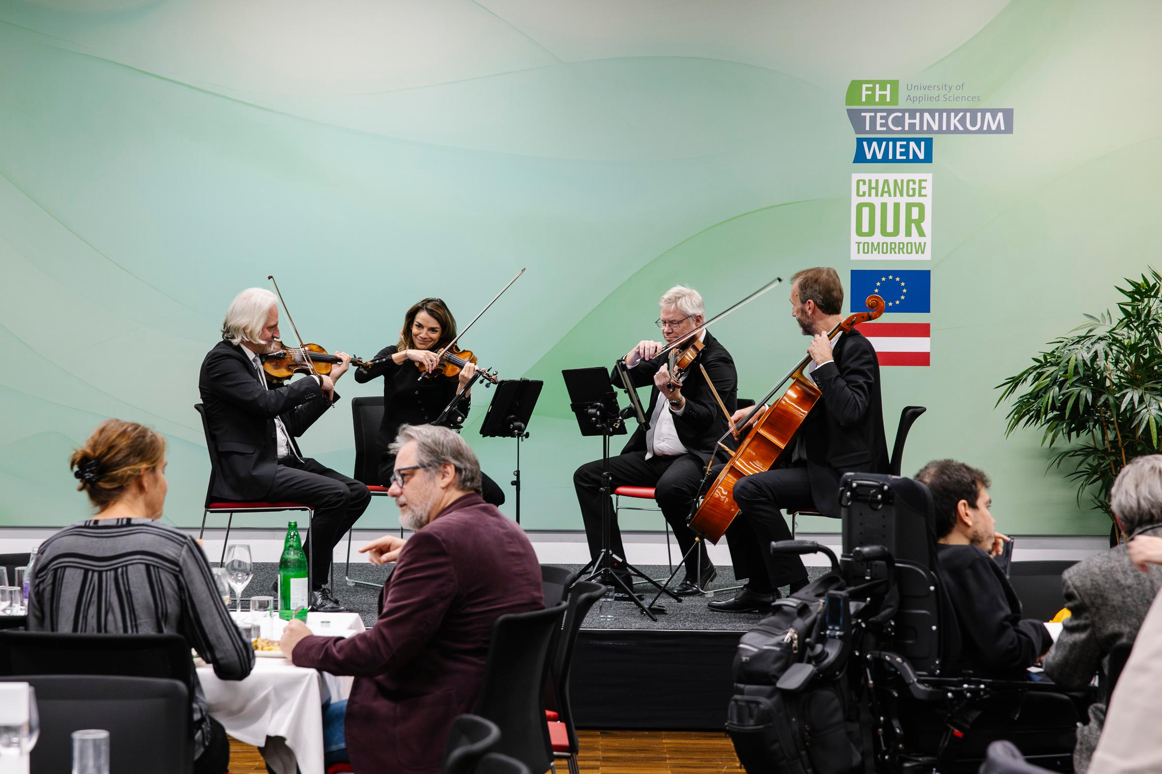 Ein Streichquartett, bestehend aus zwei Geigern, einem Bratschisten und einem Cellisten, spielt auf einer kleinen Bühne bei einer Veranstaltung. Die Gäste sitzen an Tischen im Vordergrund. Ein Transparent hinter den Musikern zeigt die FH Technikum Wien und die österreichische Flagge.