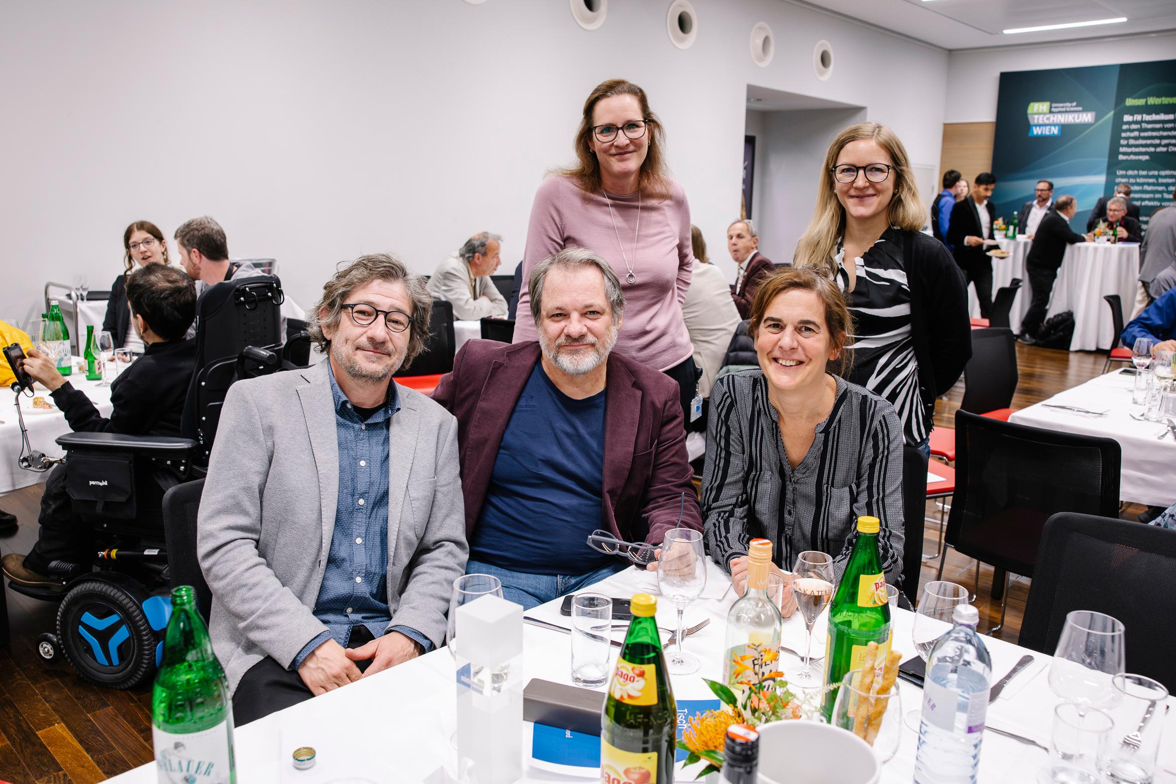 Eine Gruppe von fünf Erwachsenen, von denen drei an einem Tisch sitzen und zwei hinter ihnen stehen, lächelt für ein Foto bei einer formellen Veranstaltung, bei der Speisen und Getränke auf dem Tisch stehen; andere Teilnehmer sind im Hintergrund zu sehen.